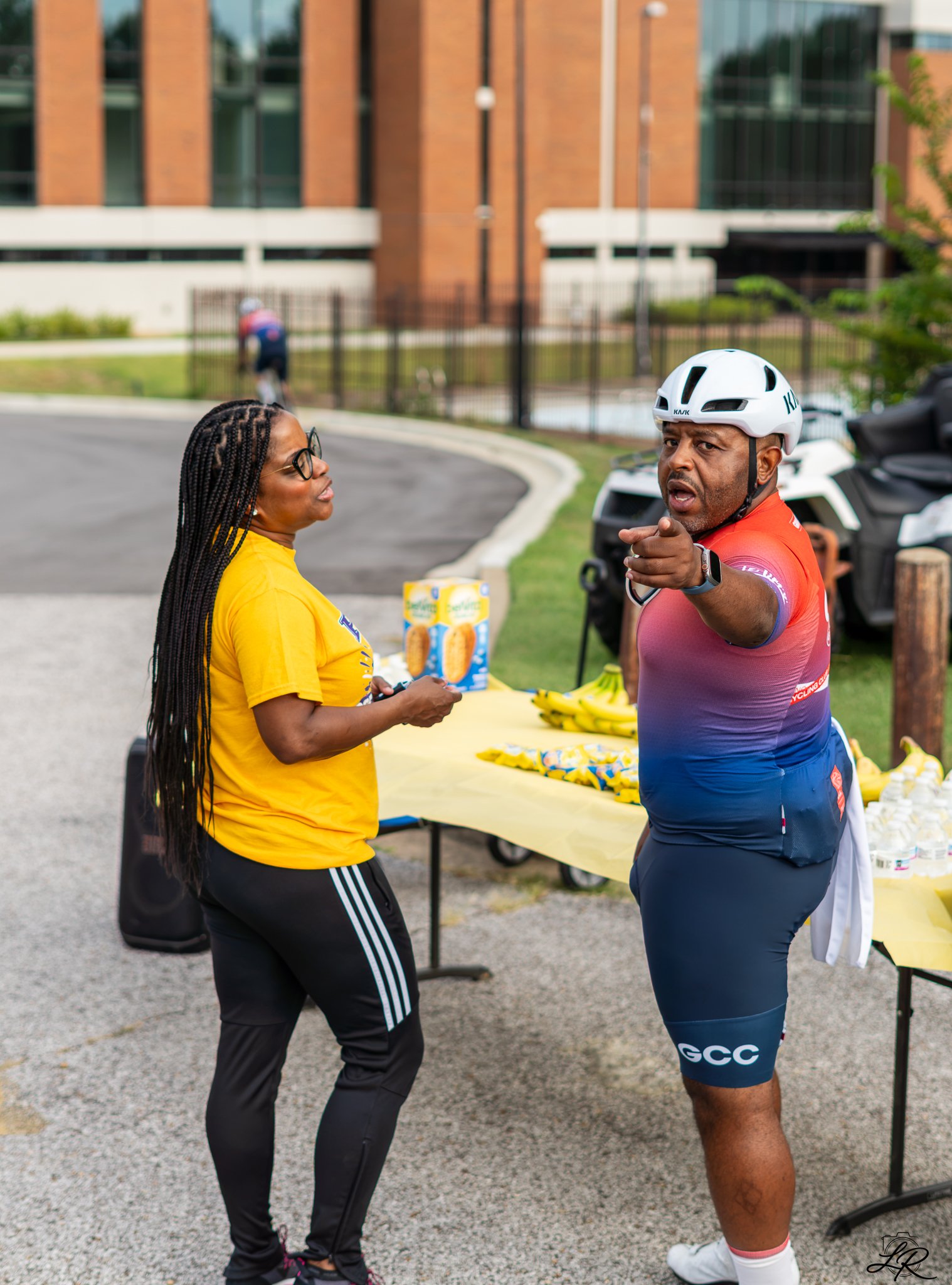 A woman in a yellow shirt and black pants is talking to a man in cycling gear, including a white helmet, blue and red shorts, and a purple and pink cycling jersey, near a table with bananas and snacks on it. They are outdoors with a building in the b