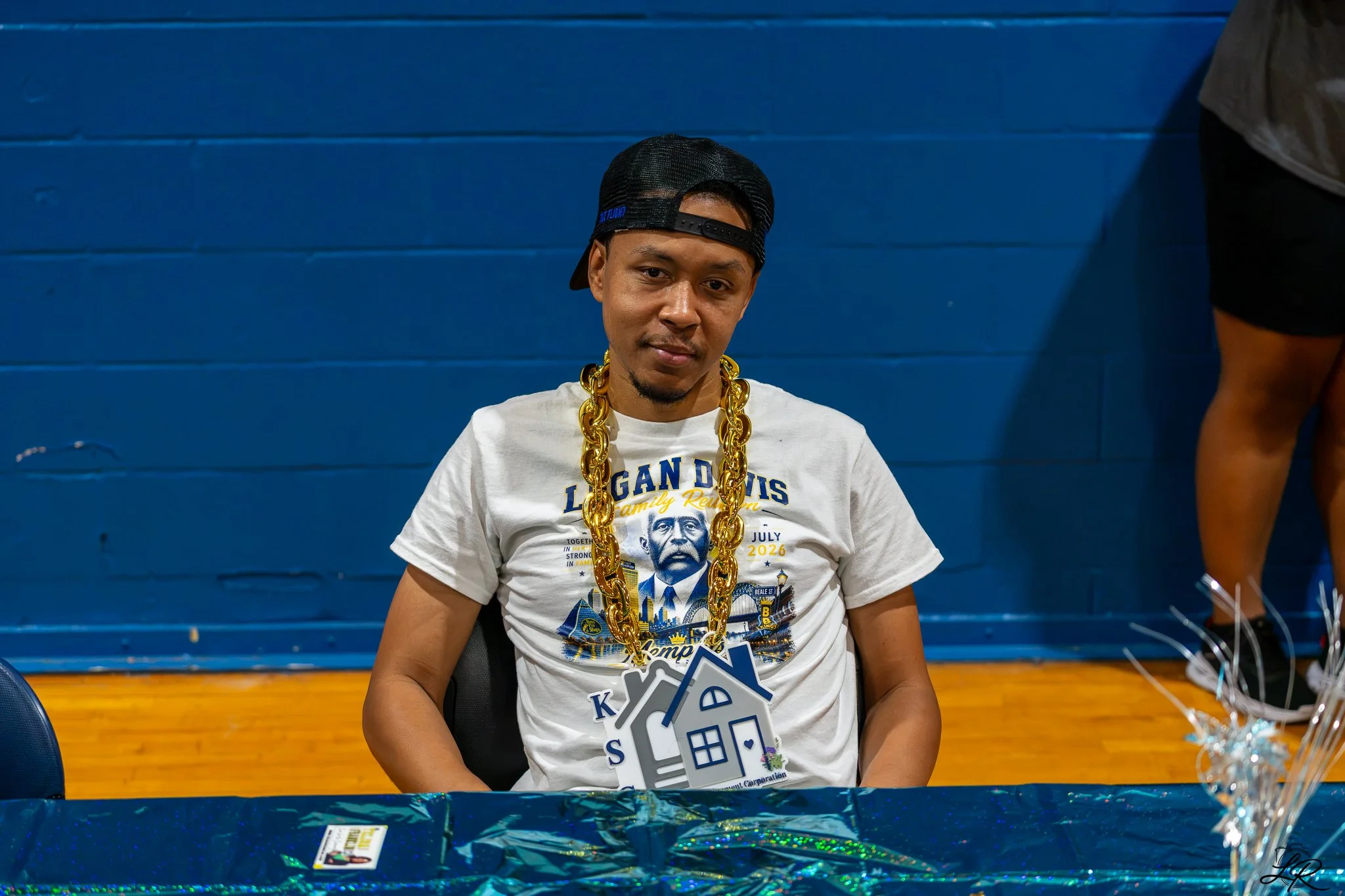 Young man sitting at a table wearing a black cap backward, a white t-shirt with a graphic design, and a large gold chain necklace. He is indoors with a blue wall behind him.