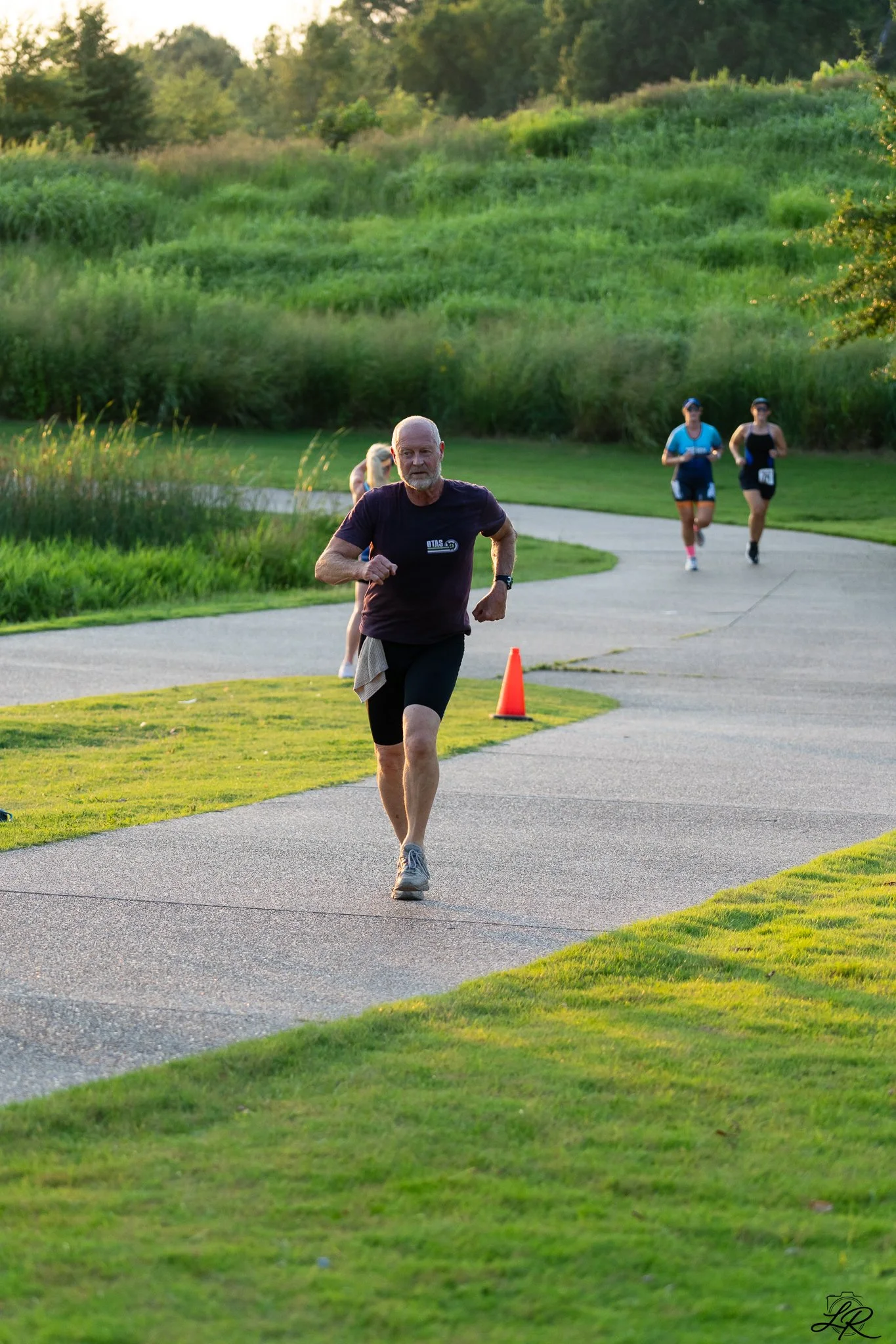 An older man with a beard jogging on a paved path in a park, with two women jogging behind him. The park has lush green grass and trees in the background, and a small orange cone is visible on the path.