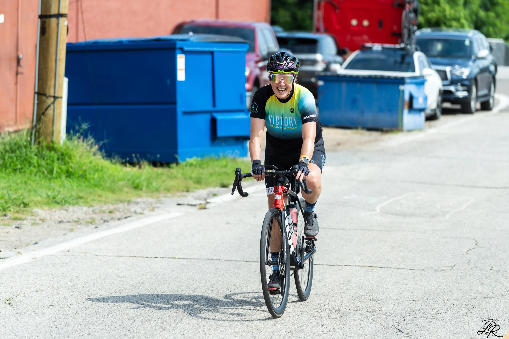 A woman riding a road bicycle on a street, wearing sunglasses, a helmet, and a cycling jersey that reads 'Victory Training Team.' She is smiling.