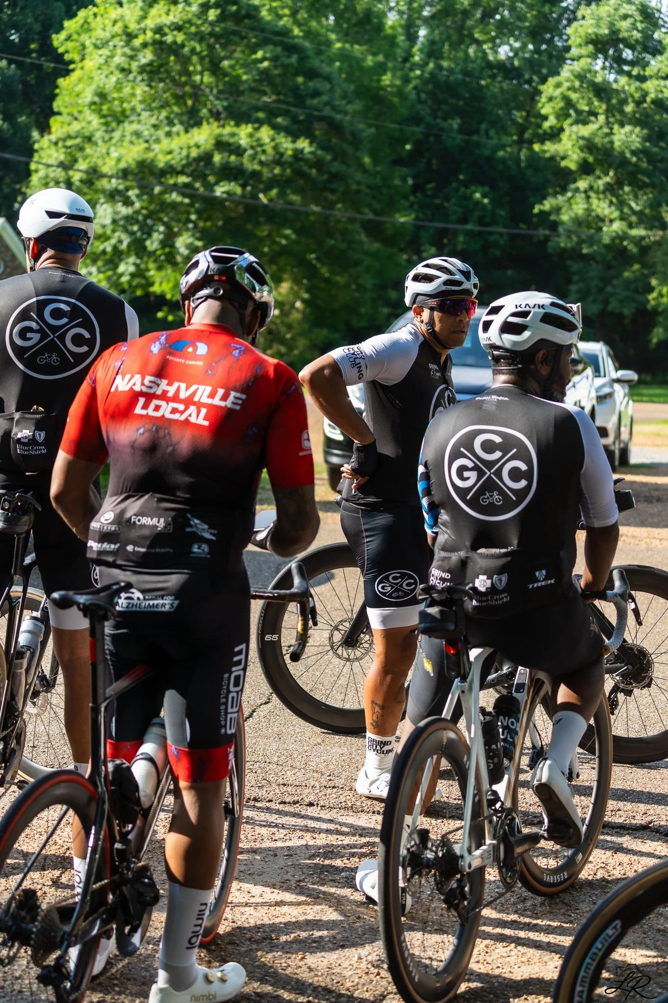 A group of cyclists gathered outdoors with lush green trees in the background, wearing helmets and cycling gear.
