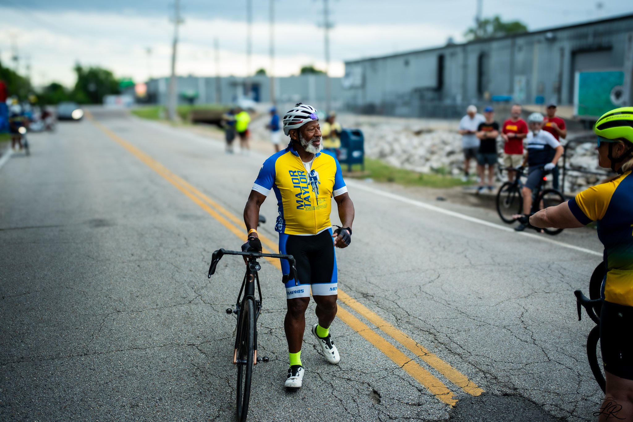 An elderly man wearing a yellow and blue cycling jersey and a white helmet stands with a black bicycle on an asphalt road with yellow dividing lines, surrounded by other cyclists and onlookers during a cycling event.