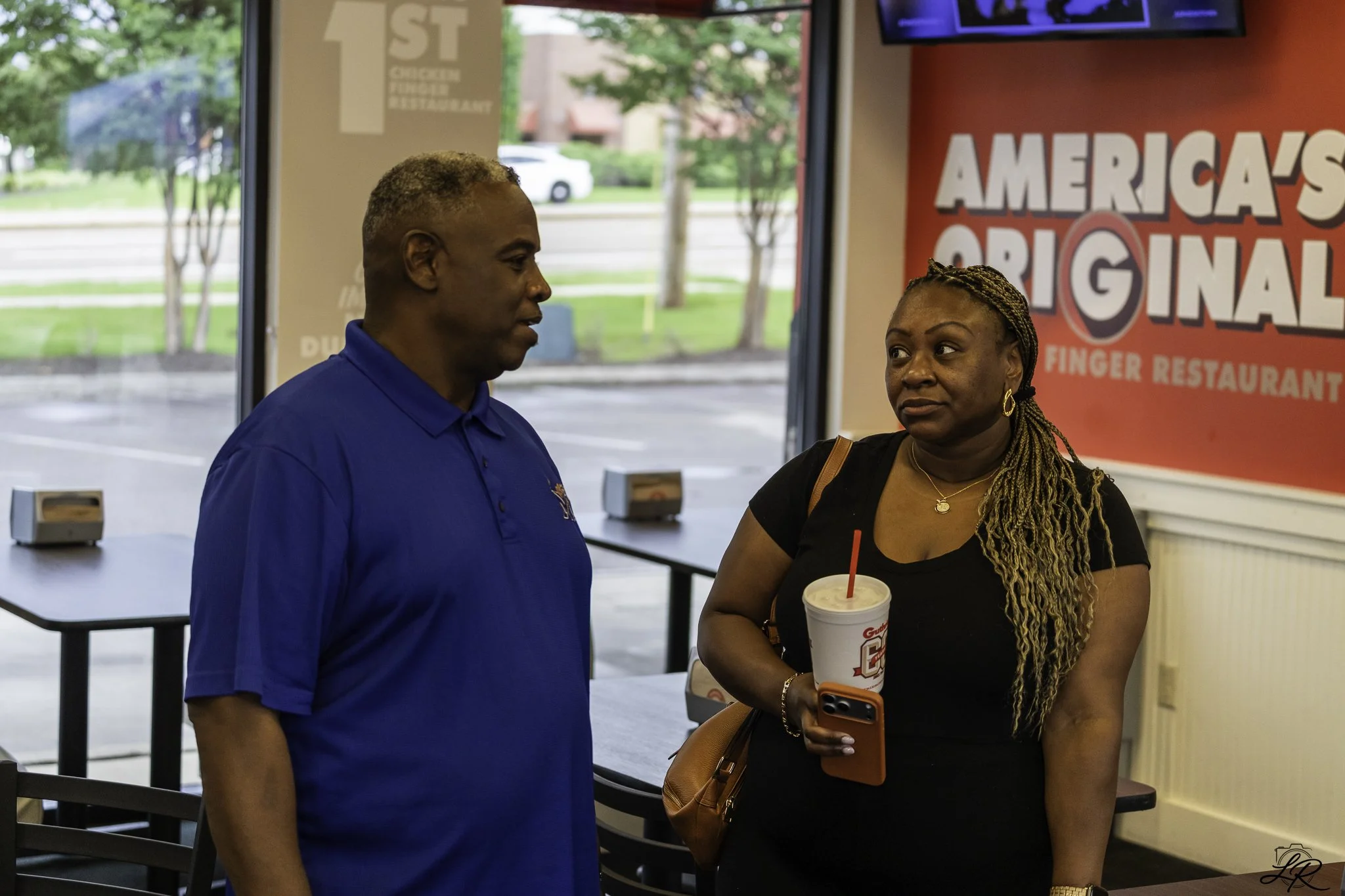 A man and a woman inside a fast-food restaurant, standing and talking to each other. The woman is holding a large drink cup with a straw and a phone. Behind them, there is a sign that says 'America's Original Finger Restaurant'.