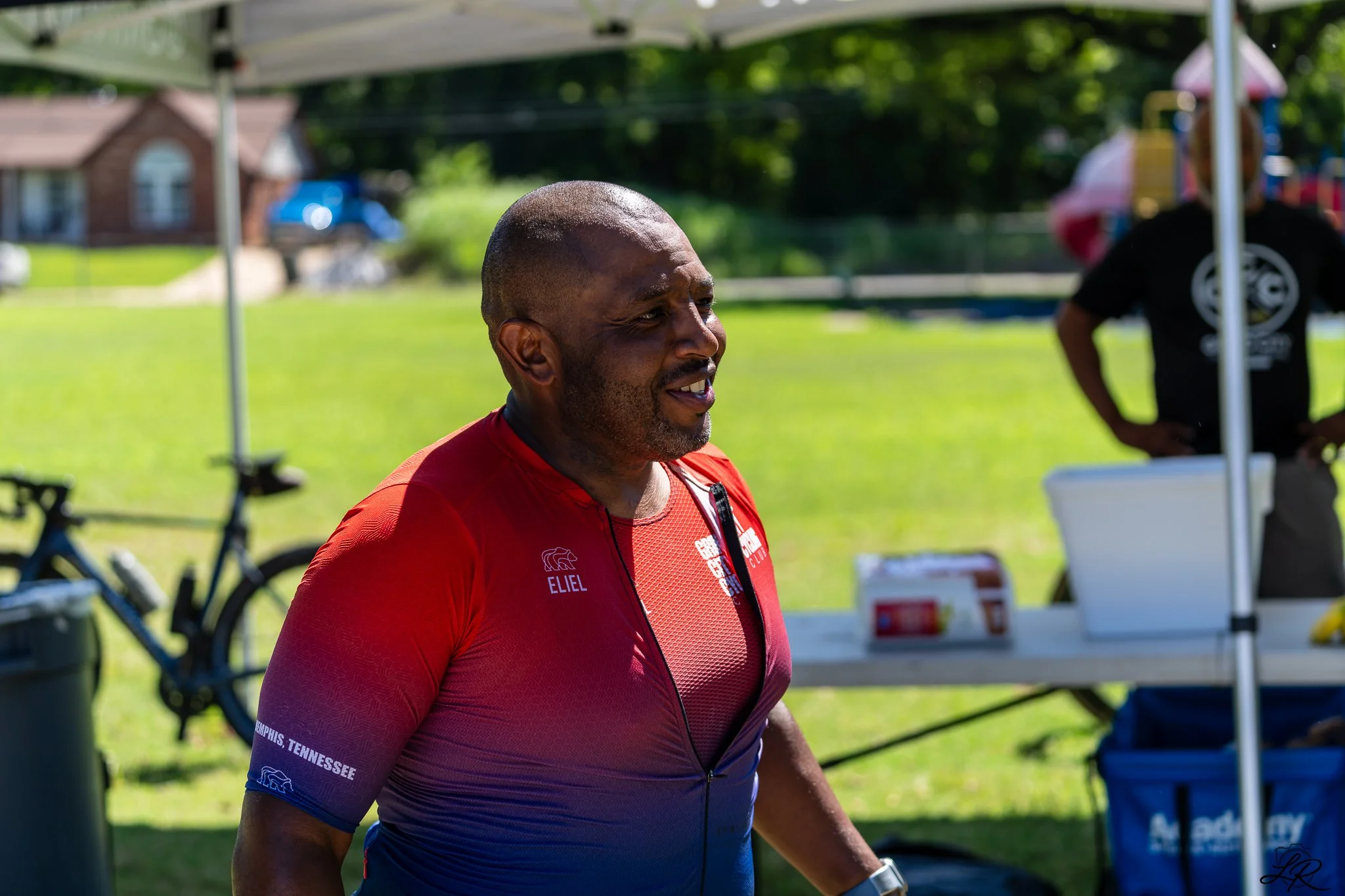 A man in a red athletic shirt outdoors with trees and a park in the background, standing near a table under a shaded canopy.