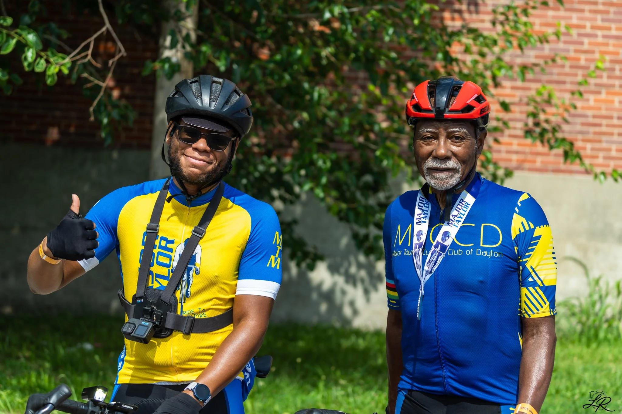 Two men wearing cycling jerseys and helmets standing outdoors near greenery and a brick wall. One man is giving a thumbs-up.