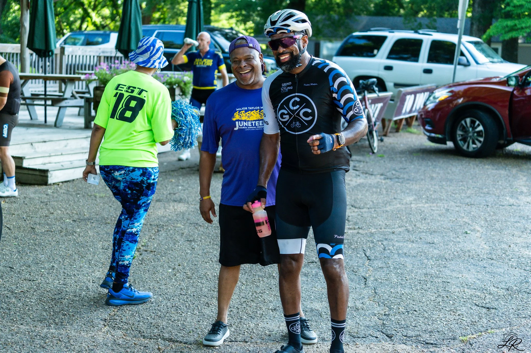 Group of people, including a man in cycling gear, smiling and talking outdoors near parked cars and trees.