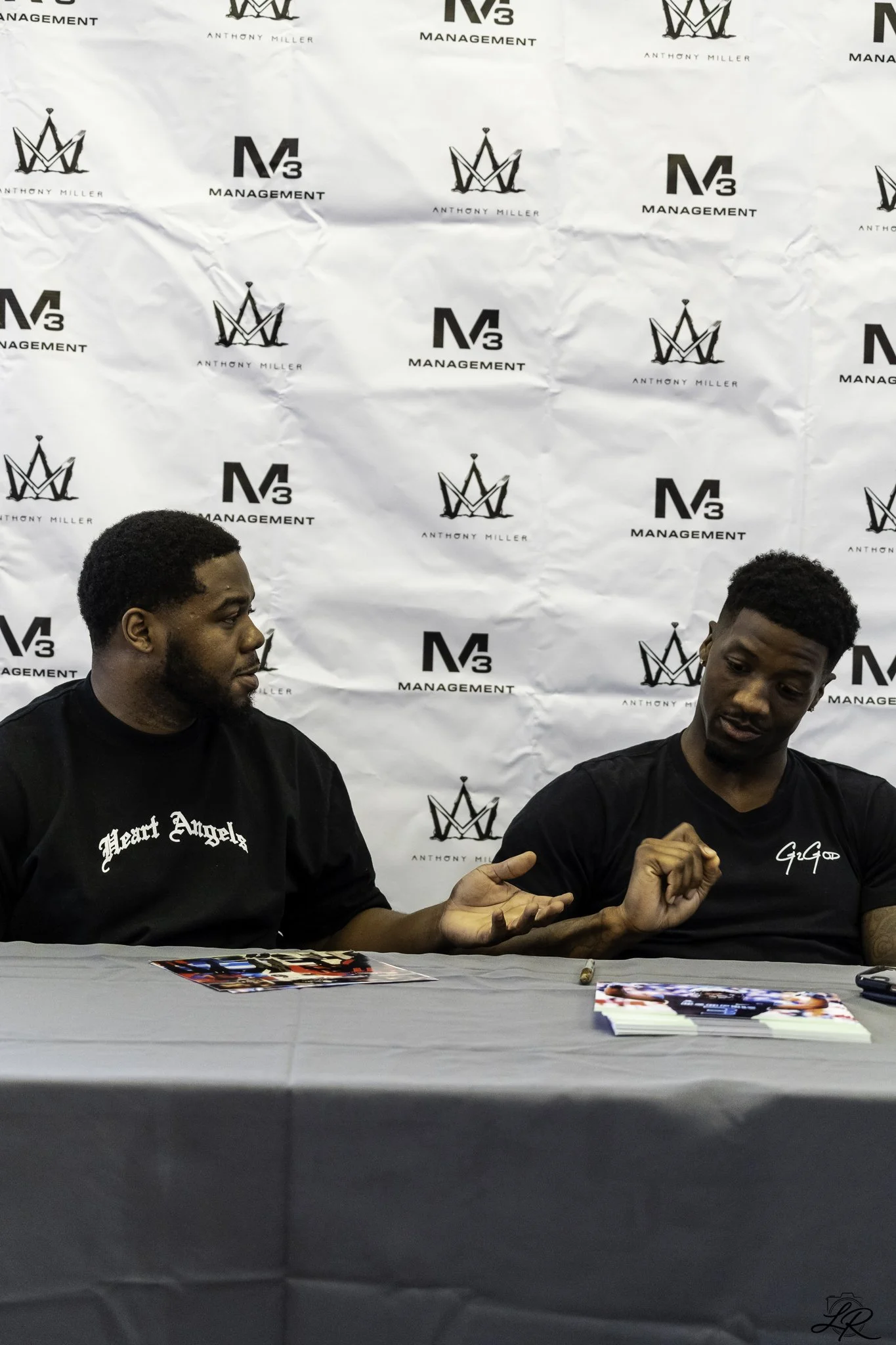 Two men seated at a table with a white backdrop displaying the logos for 'Anthony Miller' and 'N3 Management'. The man on the left wears a black shirt with white text, and the man on the right wears a black shirt with white cursive writing. They are 