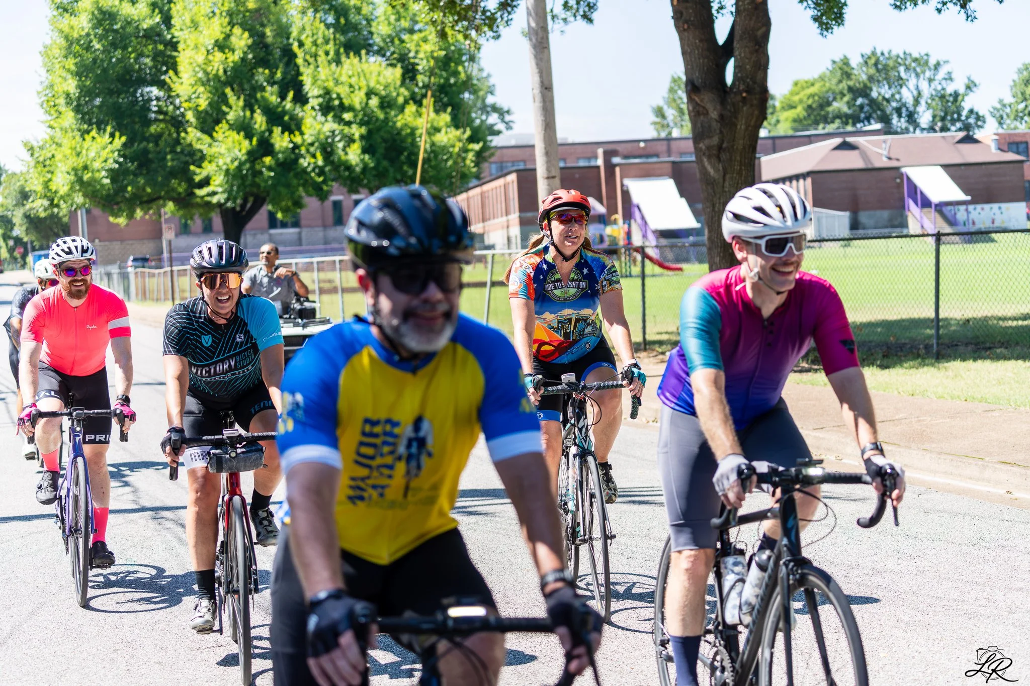 A group of cyclists riding together outdoors on a sunny day, wearing helmets and colorful athletic clothing, with trees and a playground in the background.