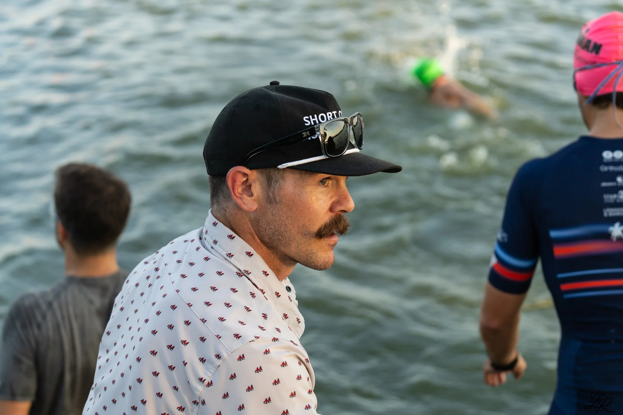 A man with a mustache wearing a white shirt with small red and black patterns and a black baseball cap with sunglasses resting on the cap, standing near a body of water, with other people in the background also in the water.