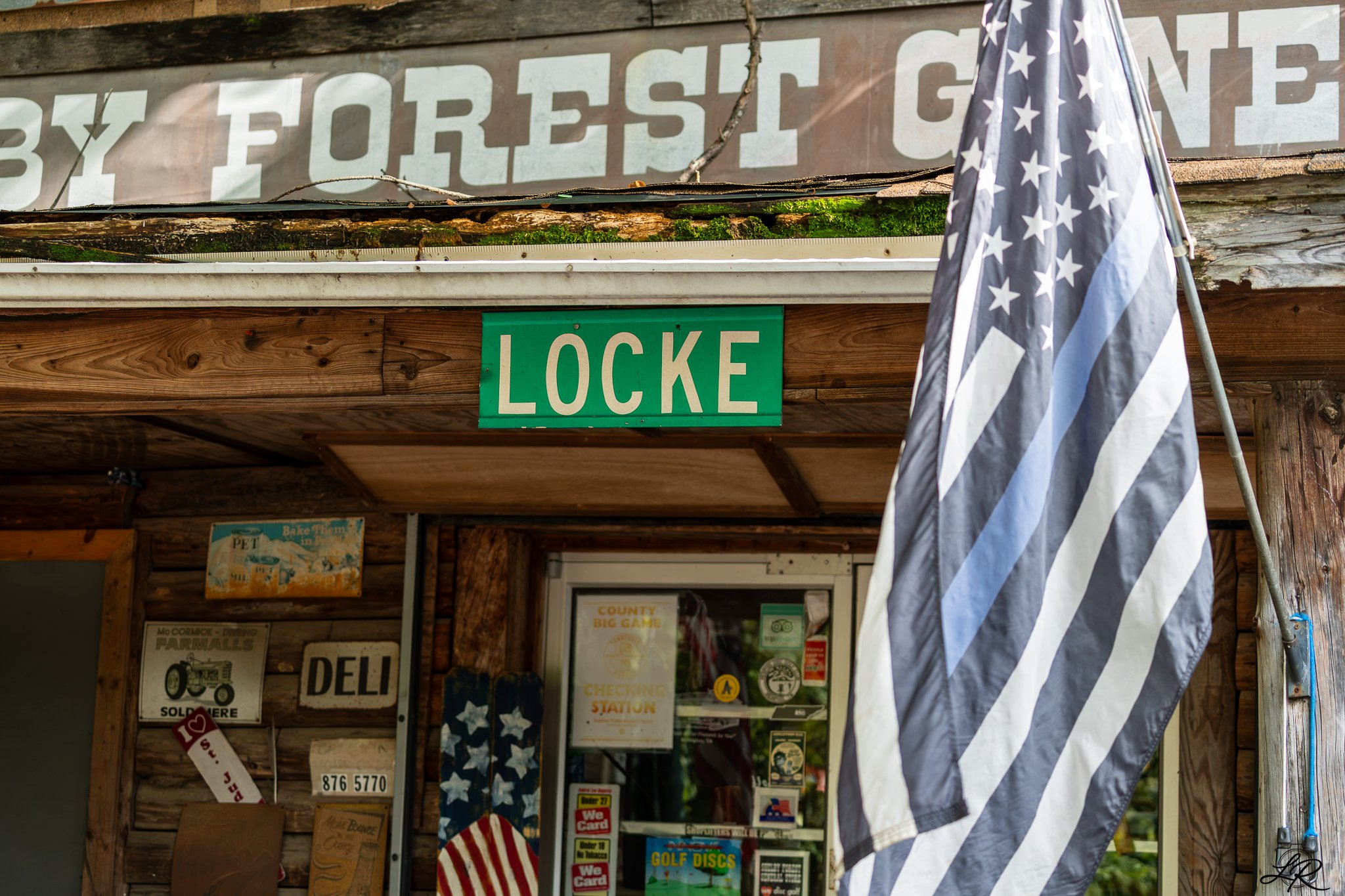 Exterior of a rustic wooden building with signs and posters, a green street sign labeled 'LOCKE', and a black and white American flag on a pole.