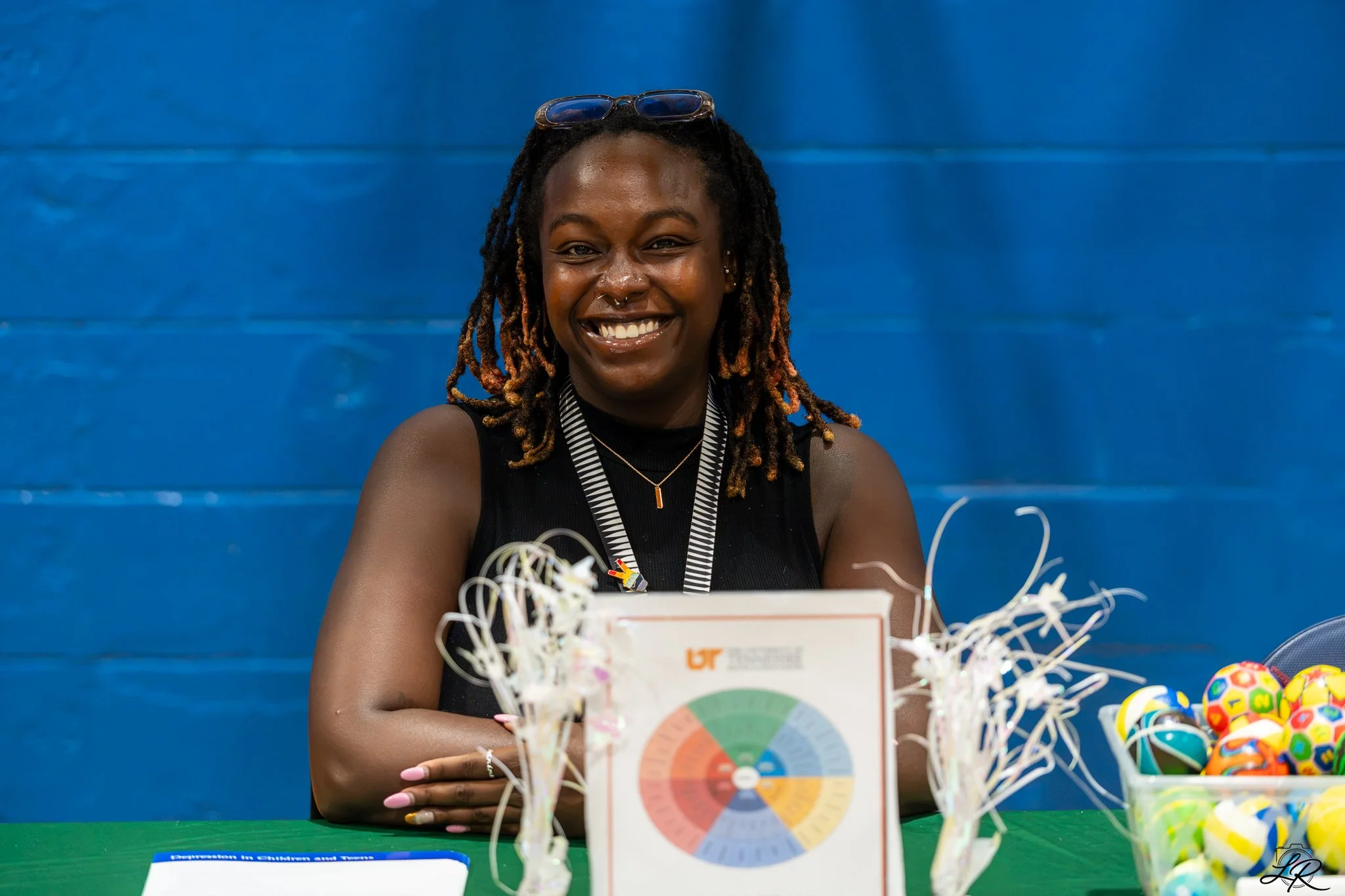 Smiling woman with glasses on her head, sitting at a table with colorful items and a chart related to children and toys, against a blue wall background.