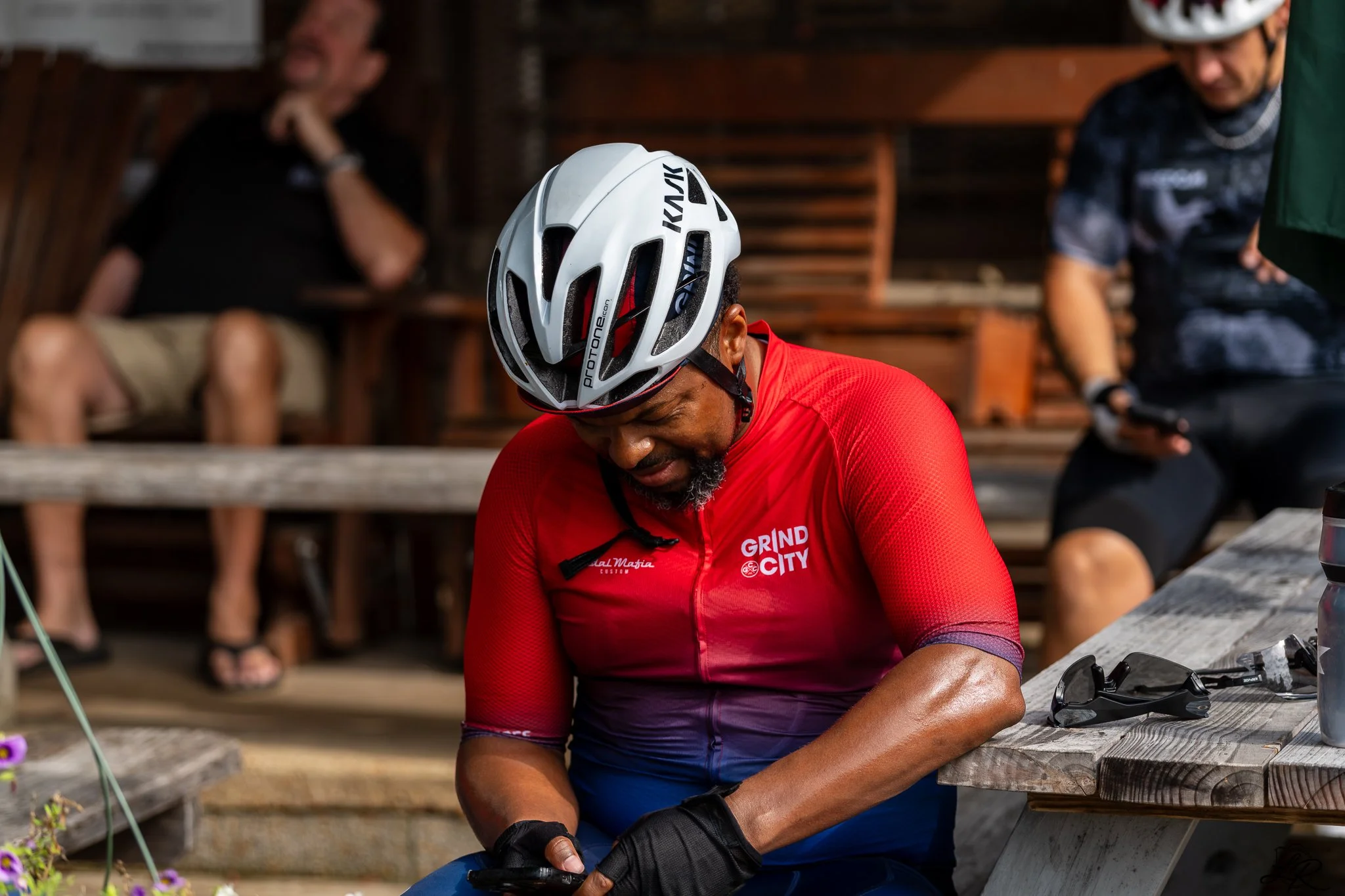 A man in a red cycling jersey and a white helmet using a phone, with two blurred people sitting on wooden benches in the background.