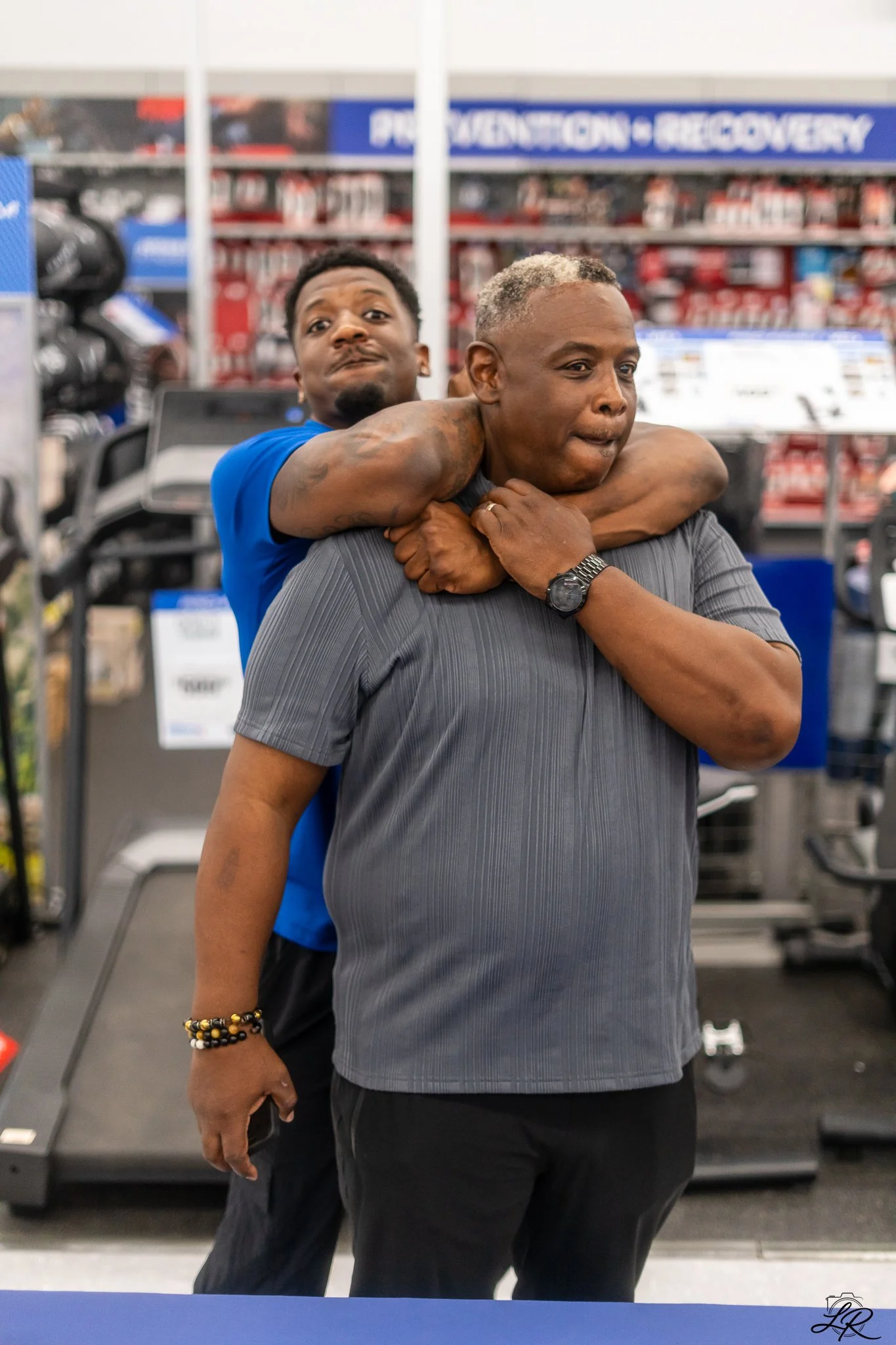 Two men in a store, one with his arms around the other in a playful pose. The background shows shelves with various products and a sign that says 'PHARMACY - RECYCLING'.