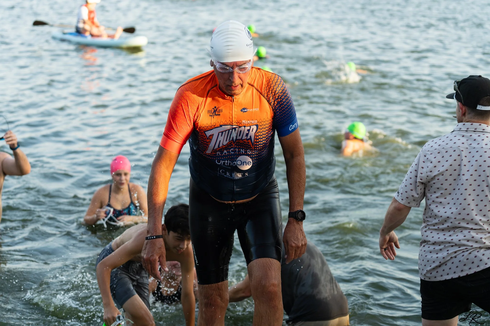 A man in an orange and blue triathlon suit and swimming cap emerges from the water during a triathlon, with other swimmers and a person on the shore visible in the background.