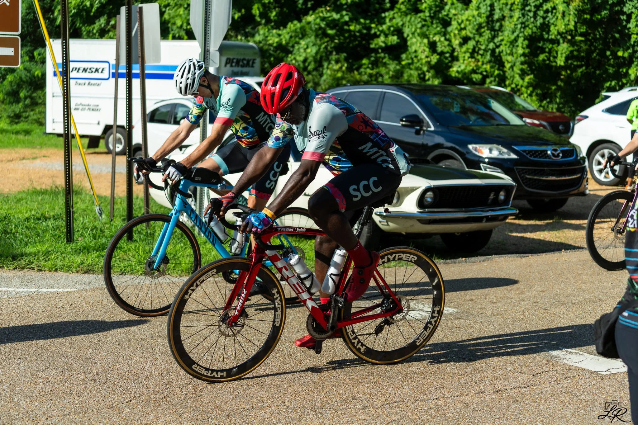 Two cyclists wearing team jerseys and helmets ride their bikes on a paved road with parked cars and green trees in the background.
