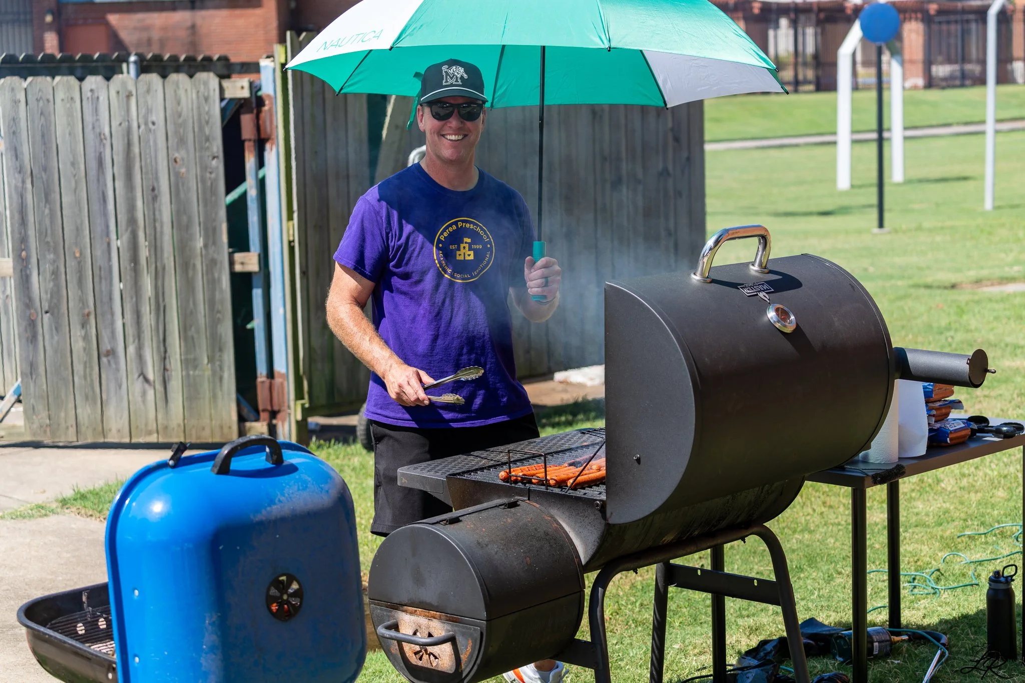 A man grilling hot dogs on a barbecue grill at an outdoor gathering, holding tongs in one hand and smiling, wearing sunglasses, a baseball cap, and a purple T-shirt with a logo.