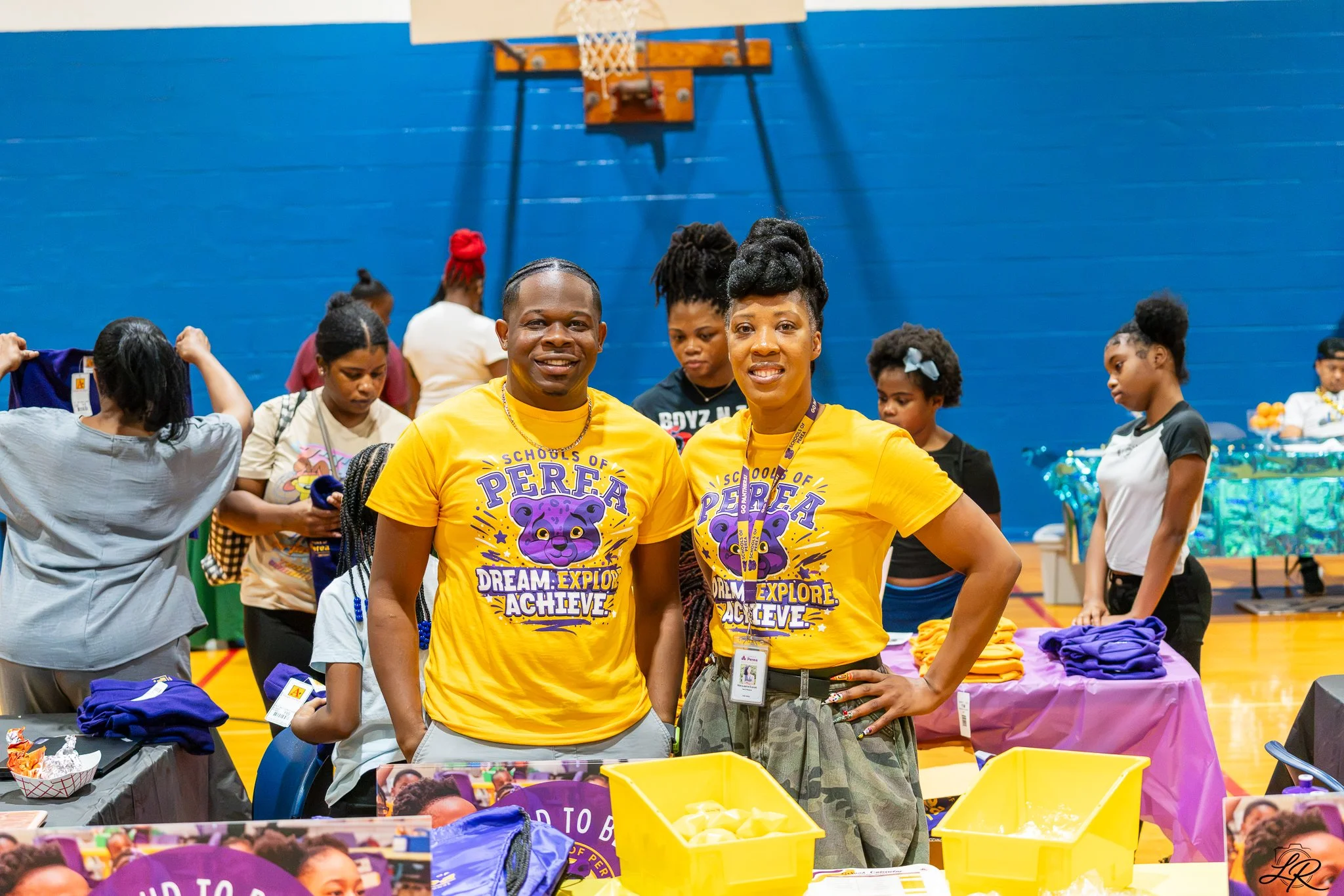 Two smiling women in yellow t-shirts with a purple lion logo and the words "Dream. Explore. Achieve." stand in the foreground of a community event inside a gymnasium. The background shows several women and girls of various ages, some looking at items