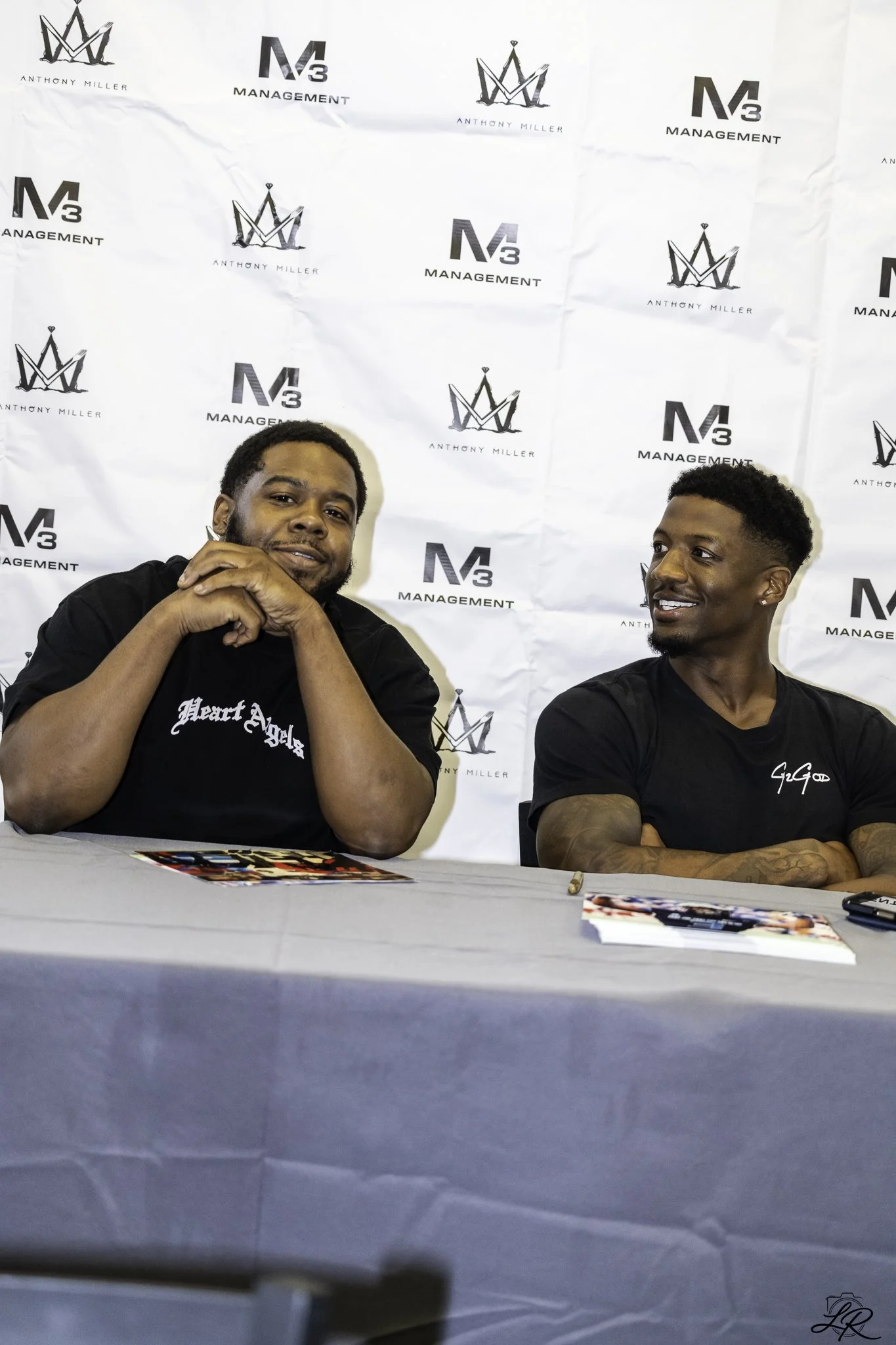 Two men seated at a table with an autograph poster behind them, two stacks of photographs on the table, and a pen in front of one man.