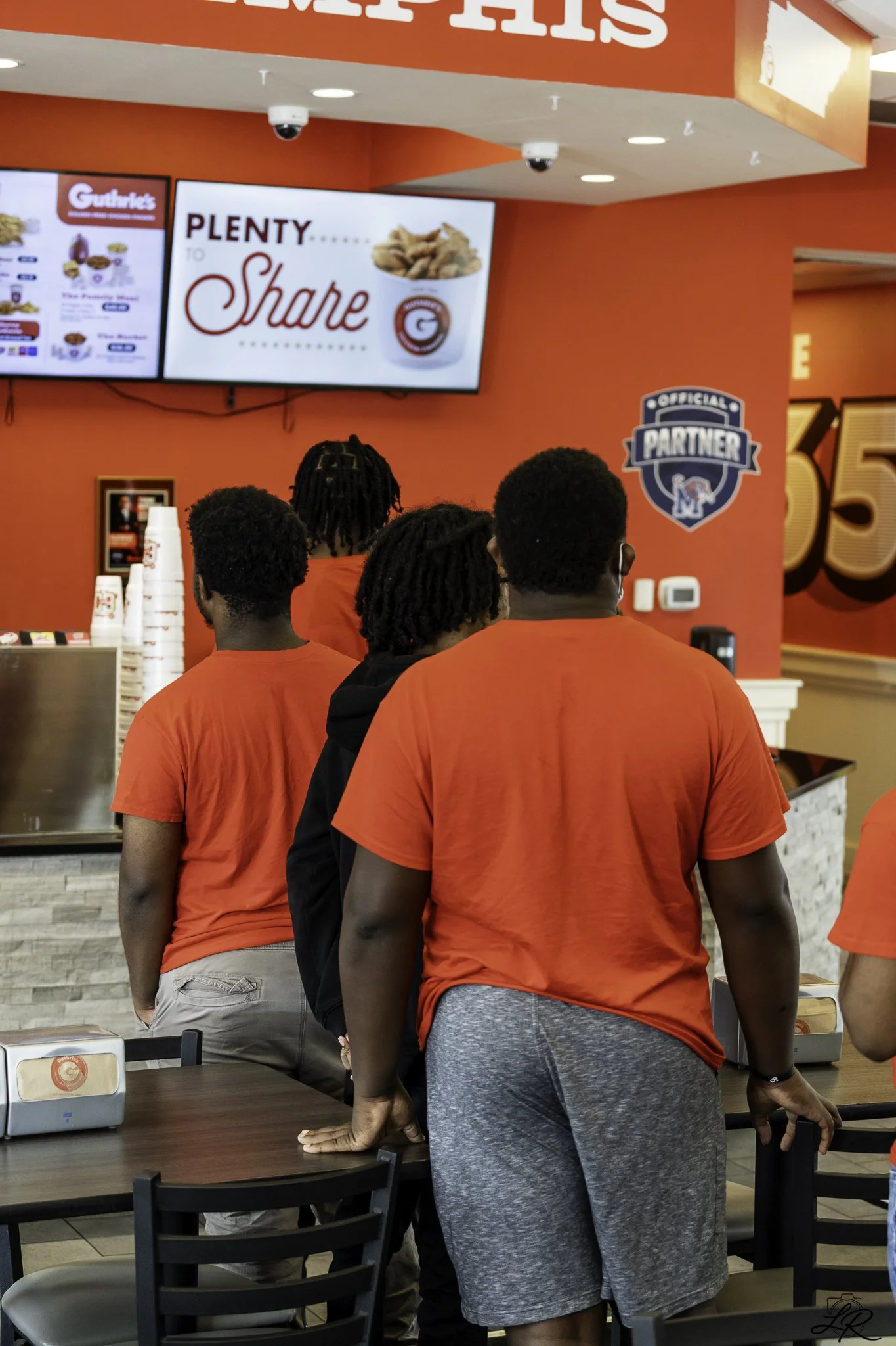 Line of people standing at a fast-food counter, facing digital menu boards inside a restaurant with red walls.