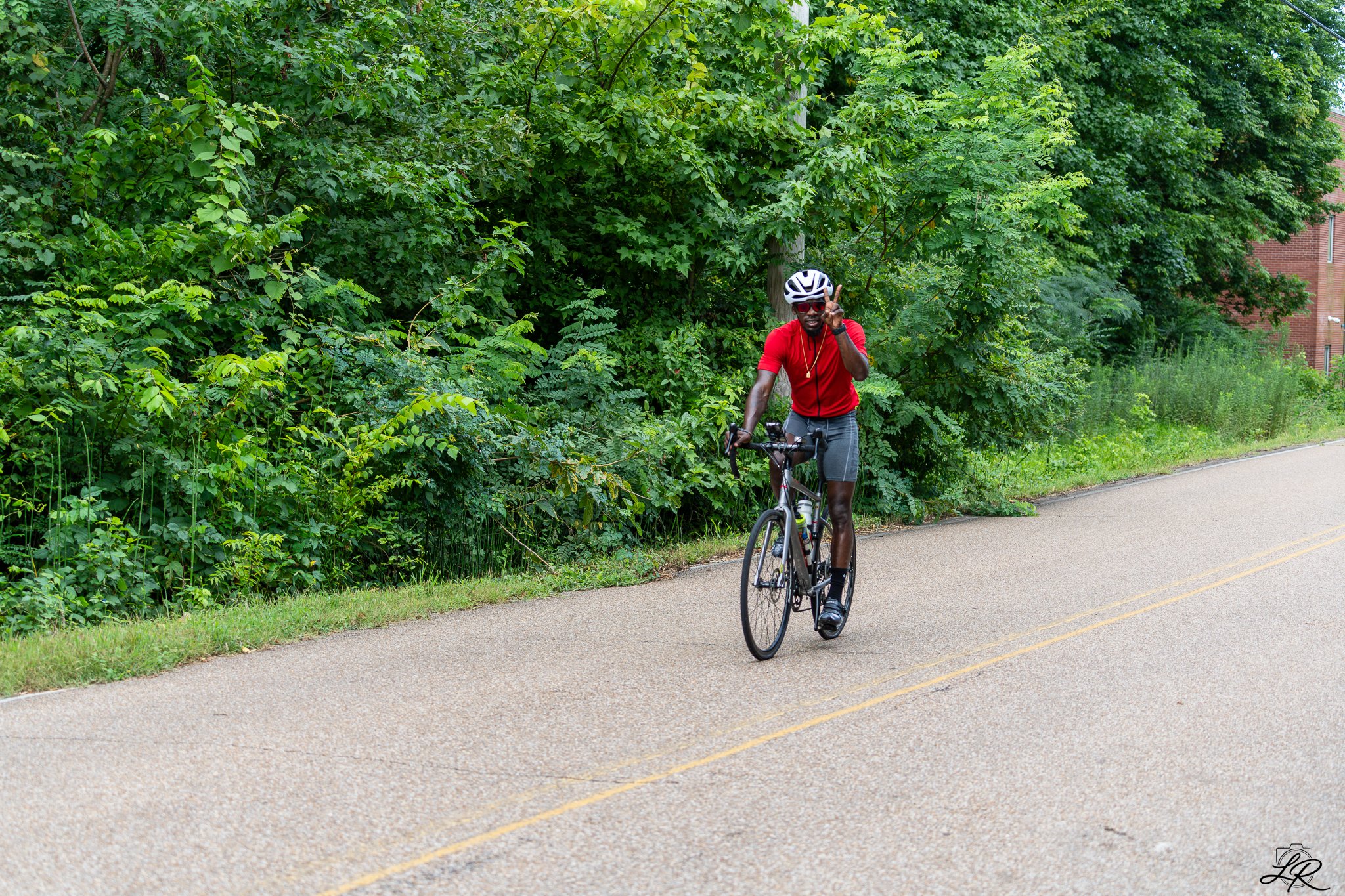 A man wearing a red shirt, shorts, and a white helmet riding a bicycle on a paved path next to dense green foliage with trees and bushes.