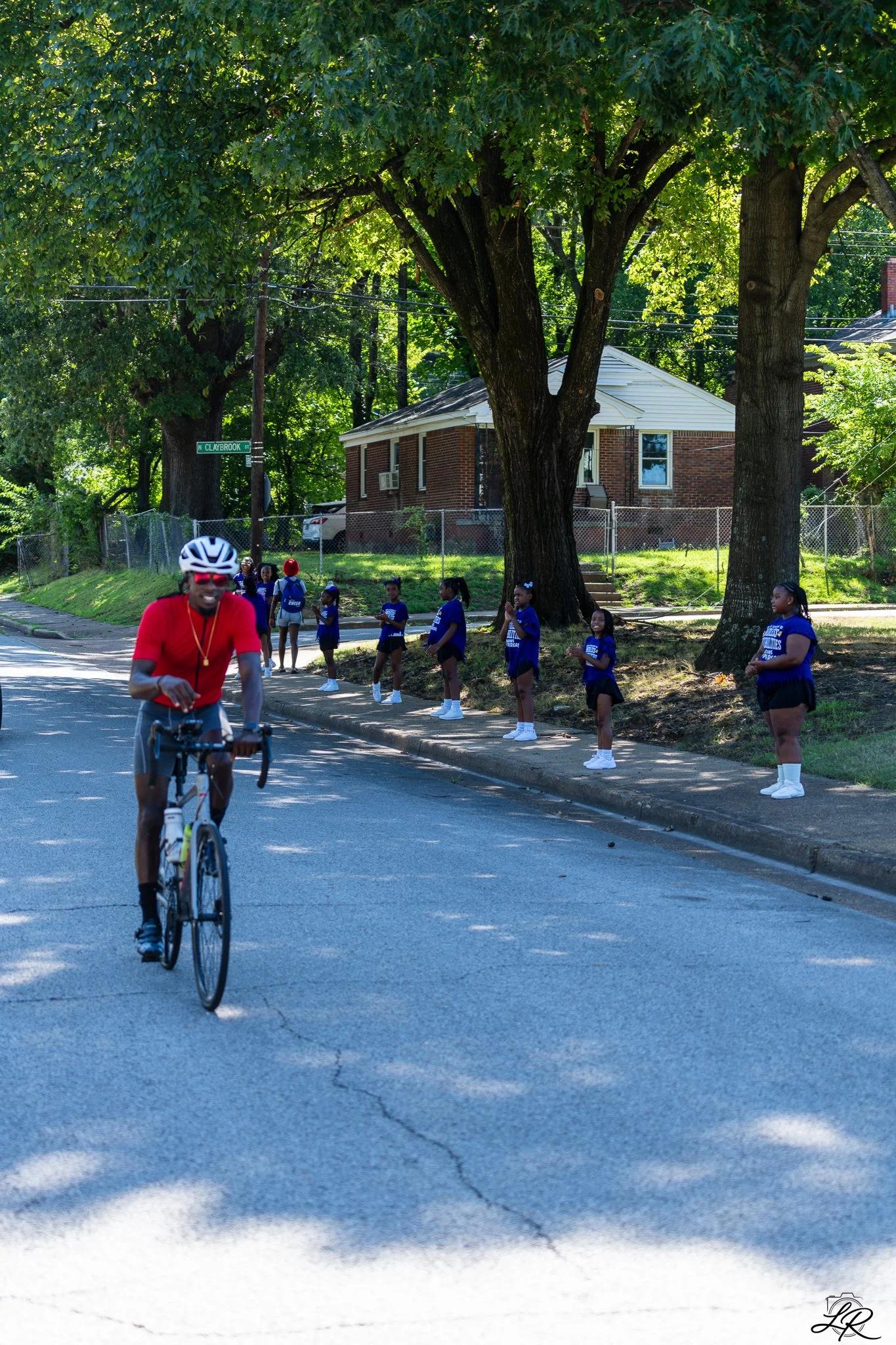 A man in a red shirt and white helmet riding a bicycle on a shaded street. In the background, a group of children in blue shirts and black skirts stand on the sidewalk, some clapping and others watching. Large trees line the street, and a brick house
