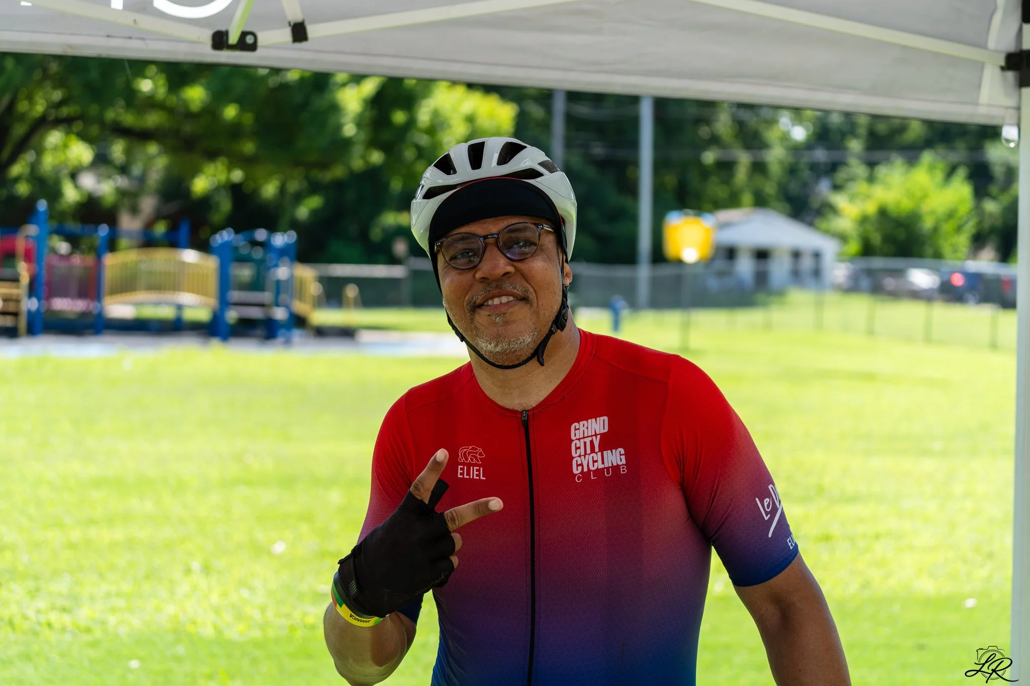 A smiling man wearing sunglasses, a white helmet, a red cycling jersey with 'Grind City Cycling Club' written on it, and a black glove is showing a peace sign with his right hand, standing under a white canopy in a park with green trees and a playgro