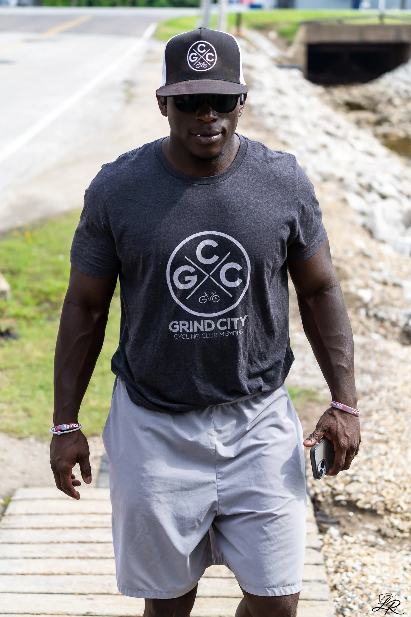 A man wearing a black cap, sunglasses, a black T-shirt with "Grind City Cycling Club Memphis" logo, gray shorts, and wristbands, standing outdoors on a stone pathway near a stream.