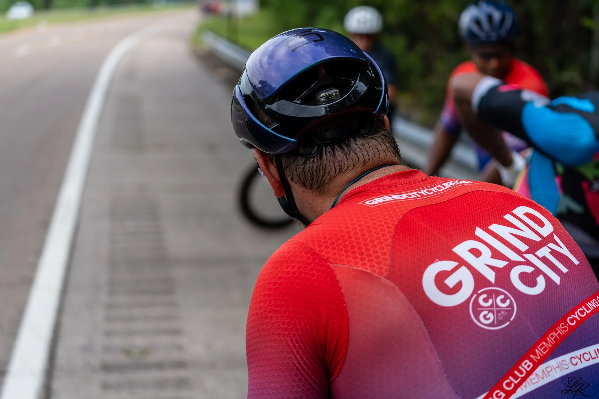 A group of cyclists wearing helmets and red jerseys, resting on the side of a road with a white dividing line, surrounded by green trees.