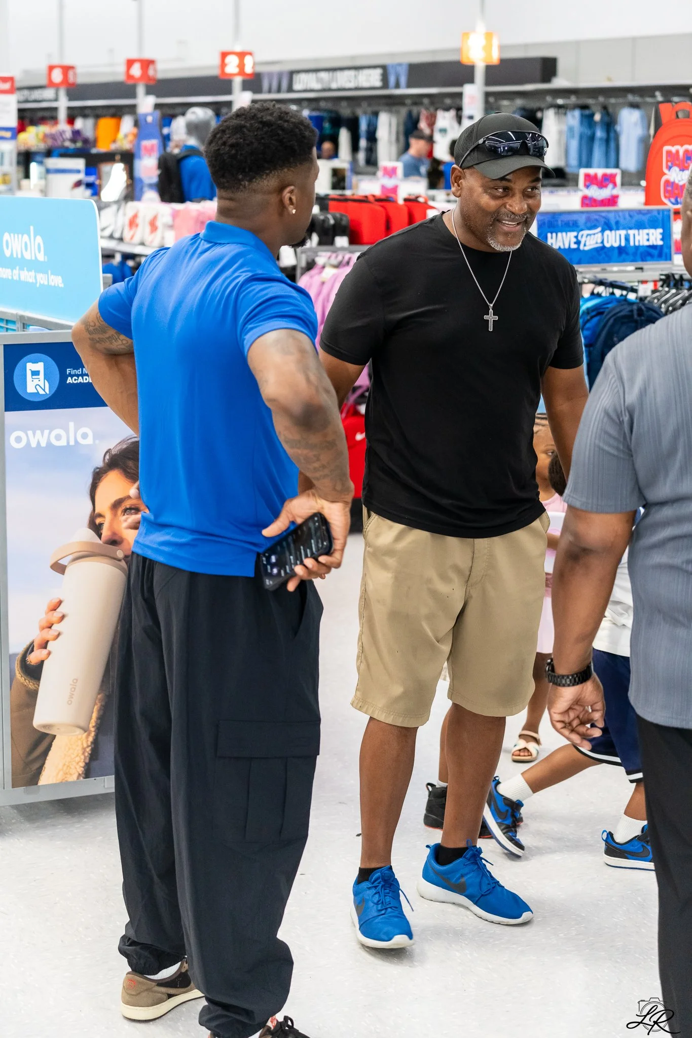 Two men talking in a retail store with clothing and shoes on display behind them. One man is wearing a black cap, black shirt, and khaki shorts, while the other has a blue shirt and black pants, holding a phone and a water bottle.