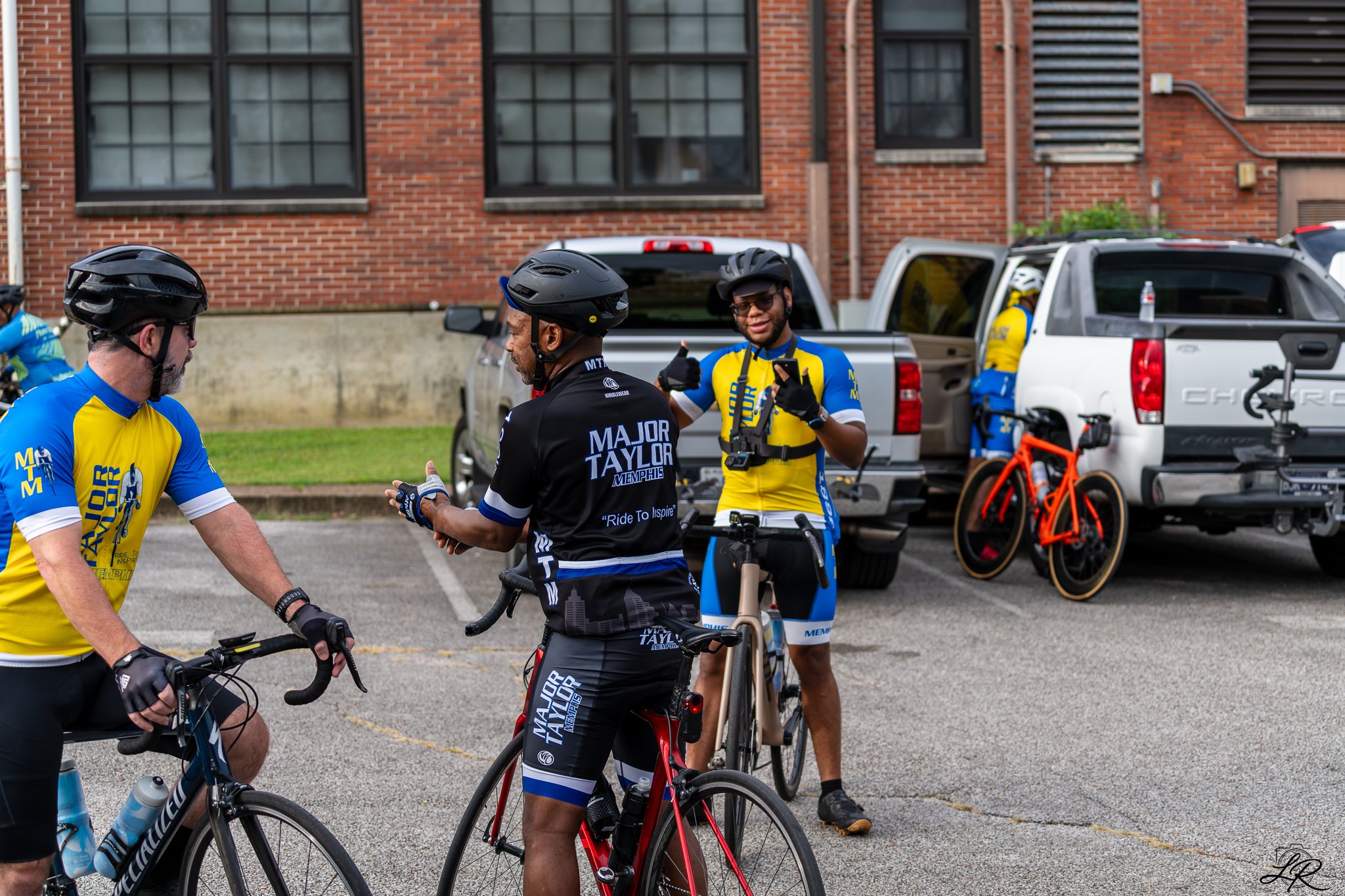 Group of cyclists in colorful jerseys and helmets, gathered near parked trucks and bicycles, engaging in conversation outdoors in front of a brick building.