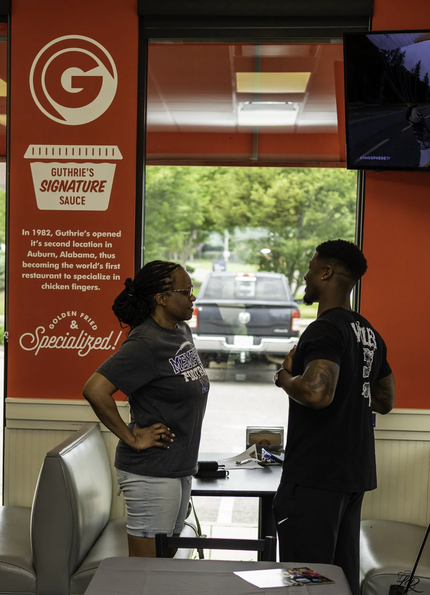 Two people standing and talking inside a restaurant or fast food place, with a red wall and a sign about Guthrie's Signature Sauce. The woman has dark hair, glasses, and is wearing a dark t-shirt and light shorts. The man has short hair, tattoos on h