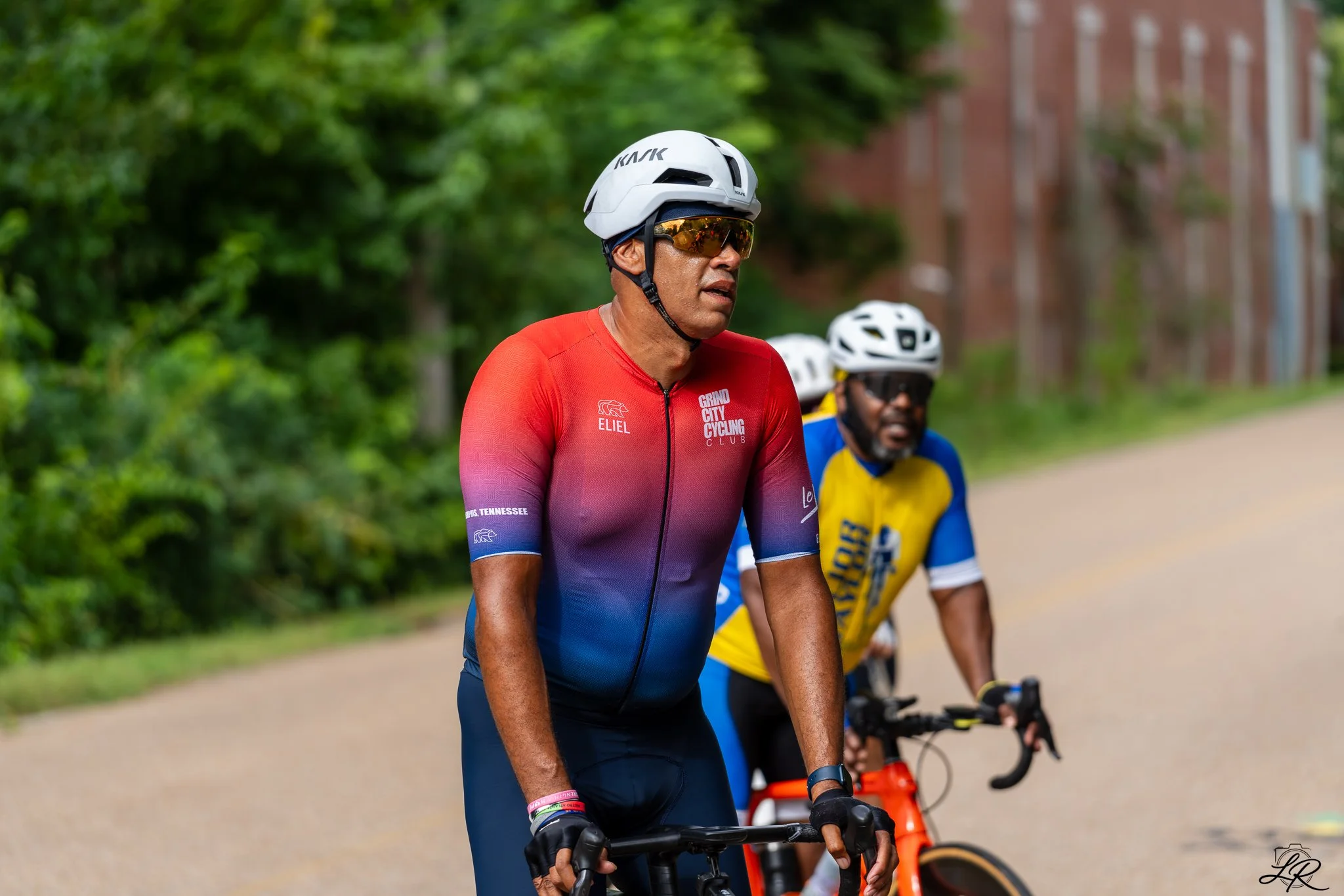 Two cyclists riding along a tree-lined pathway, wearing helmets and colorful cycling jerseys, with a brick building in the background.