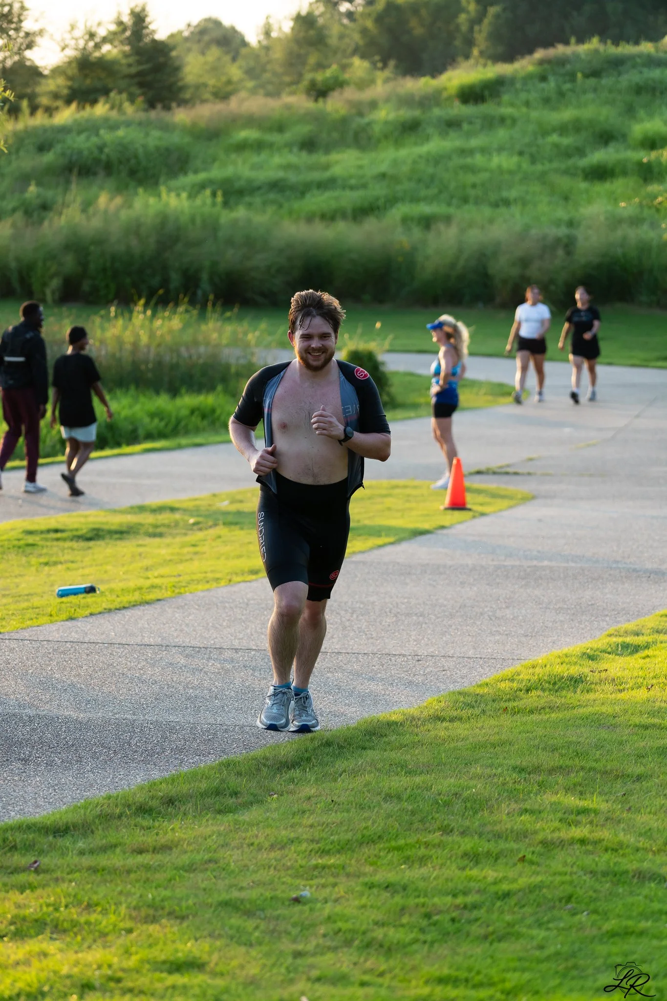 A man with a beard wearing a running vest and shoes is running outdoors on a paved path. Several other people in athletic clothing are walking or running in the background. The scene is set in a park with grassy areas, trees, and a pond, illuminated 