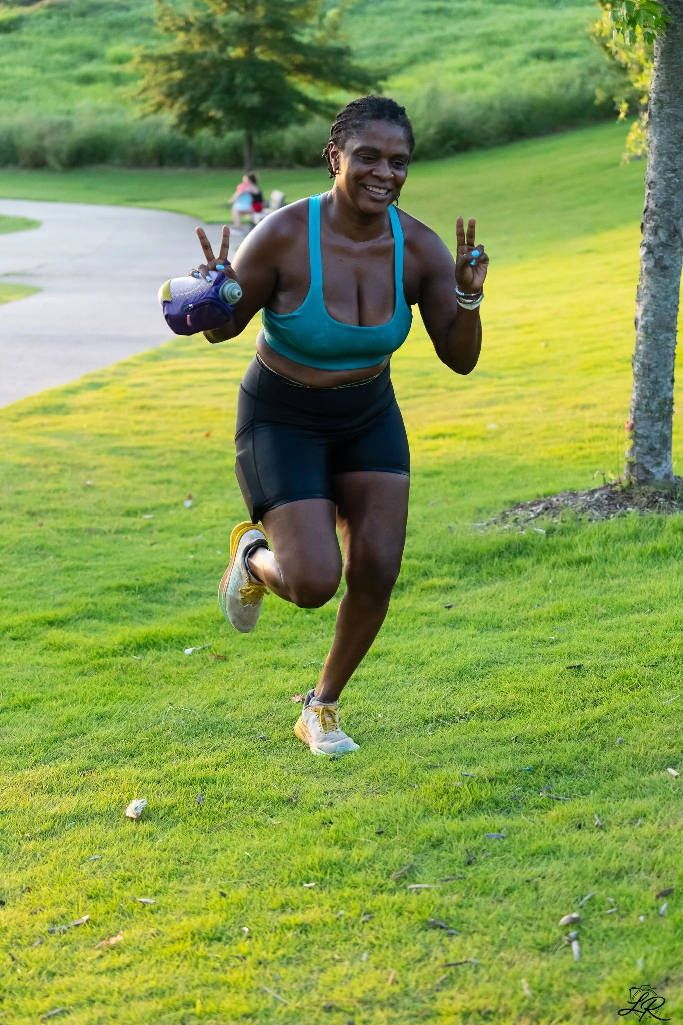 A woman running outdoors on a grassy park in the late afternoon, smiling and making peace signs with her fingers on both hands. She is wearing a teal sports top, black shorts, and yellow running shoes, with a small bag on her left arm holding a water