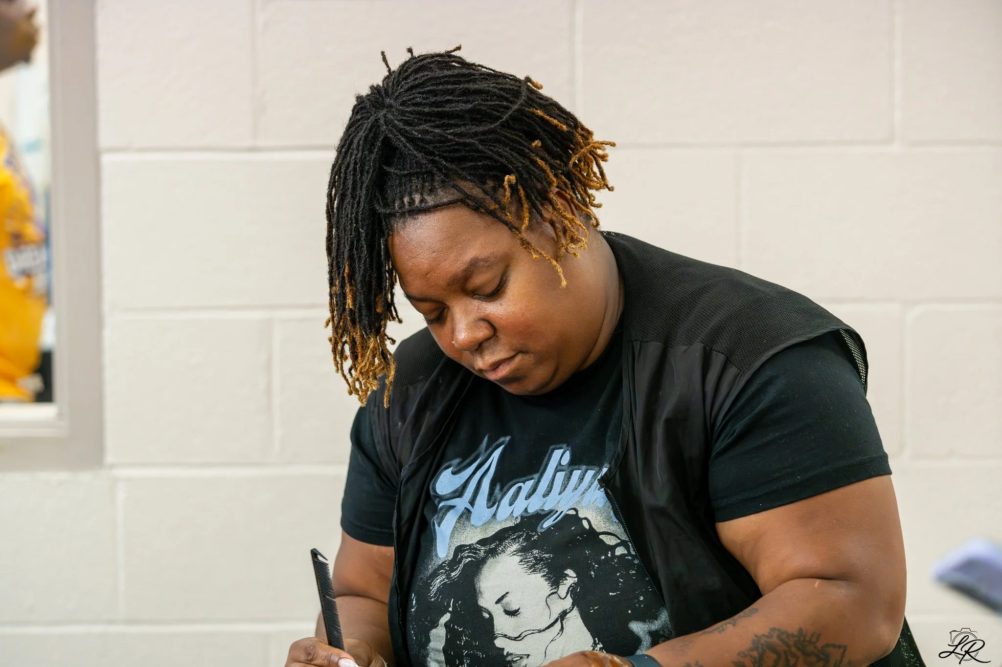 A person with dreadlocks is looking down, wearing a black Nike graphic t-shirt and appears to be seated at a table. The background features a white wall with horizontal lines.