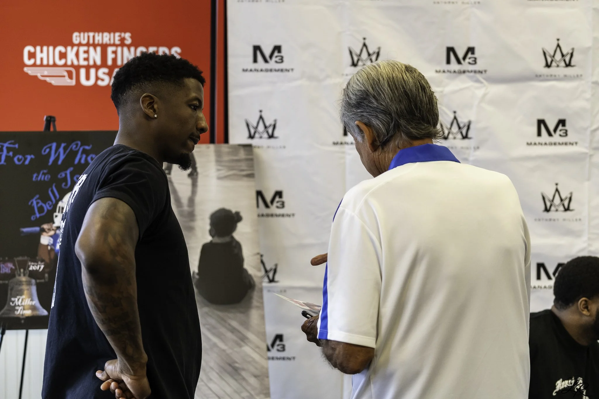 Two men are having a conversation at an indoor event, standing near a backdrop with logos. One man is dressed in a black t-shirt, and the other is in a white jacket with blue accents. There are posters and banners in the background.