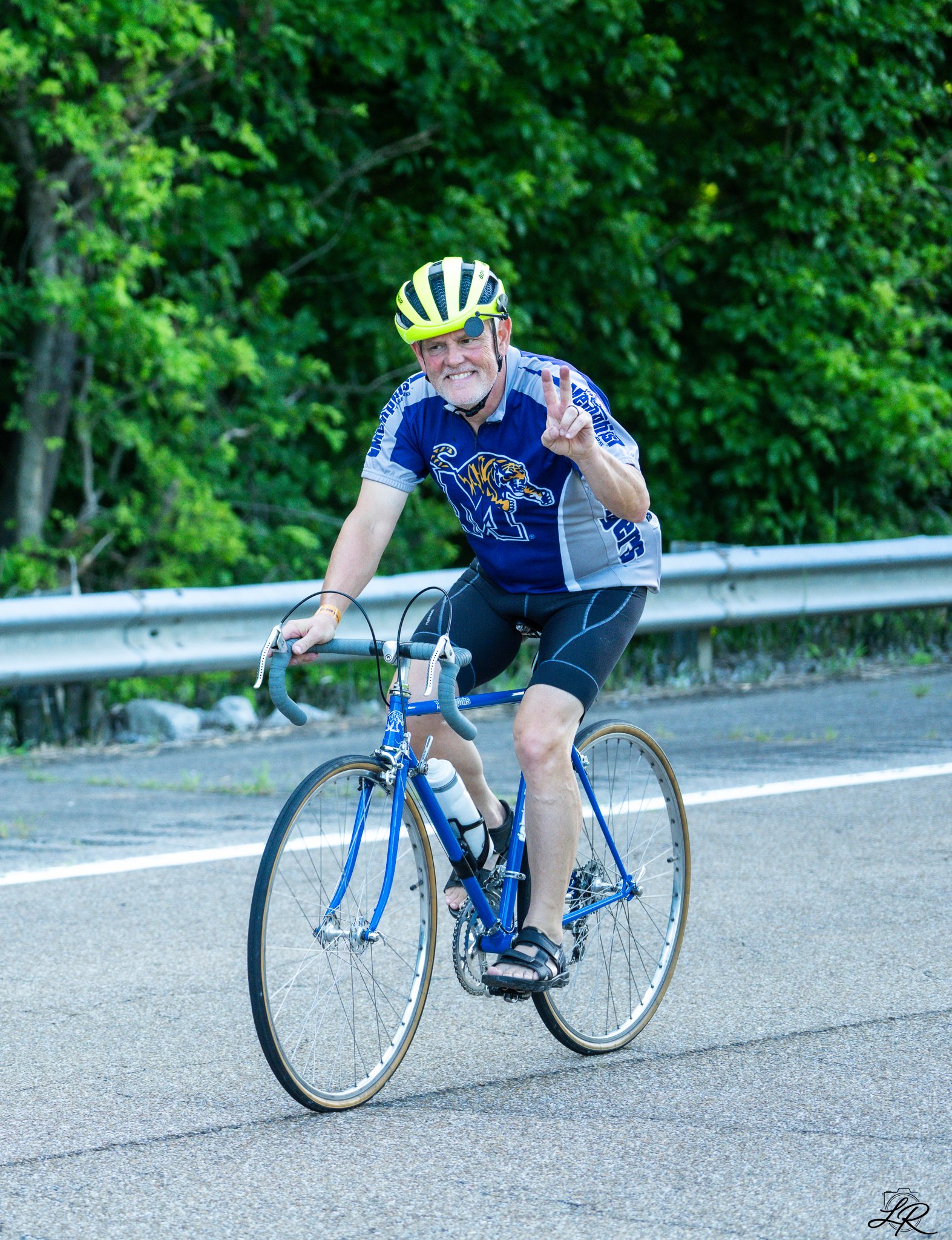 A smiling middle-aged man wearing a bright yellow bike helmet, a blue sports shirt with a mascot tiger, black shorts, and sandals, riding a blue bicycle on a paved road beside a metal guardrail with green trees in the background. He is making a peace