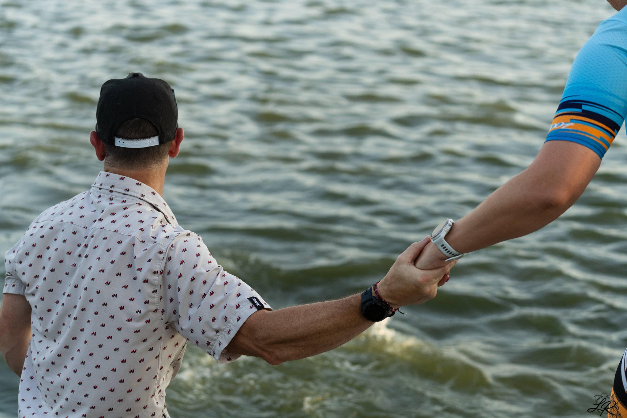 A man with a patterned white shirt and a black cap stands by the water, holding the arm of another person wearing a blue and orange patterned shirt, in a scene by a body of water.