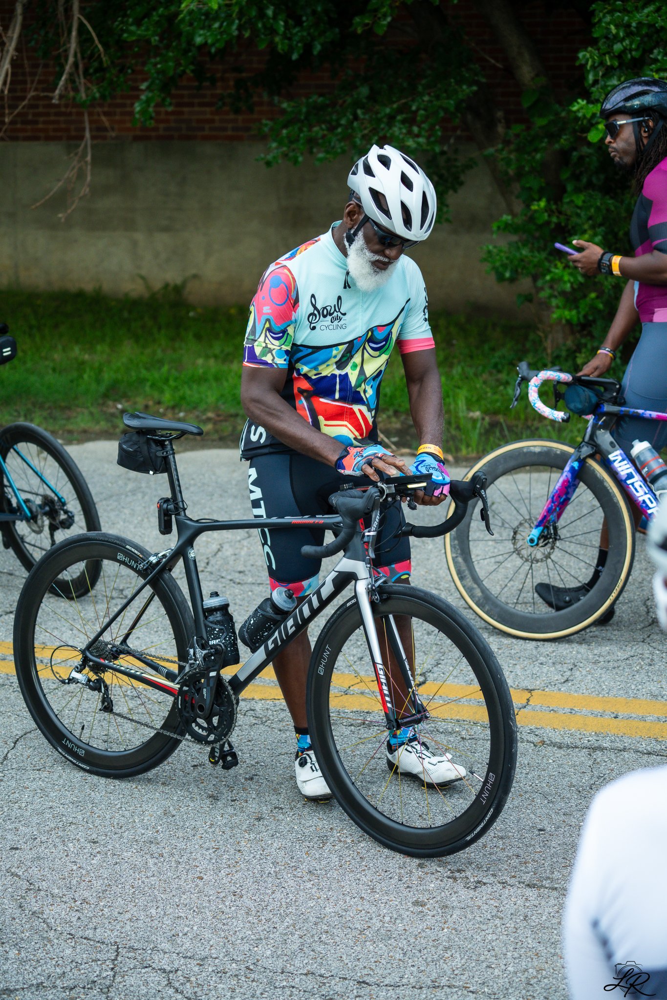 An older man with a white beard, wearing a colorful cycling jersey, black cycling shorts, a white helmet, and sunglasses, is standing next to a black bicycle. A woman in a purple and black cycling outfit, wearing a black helmet and sunglasses, is hol