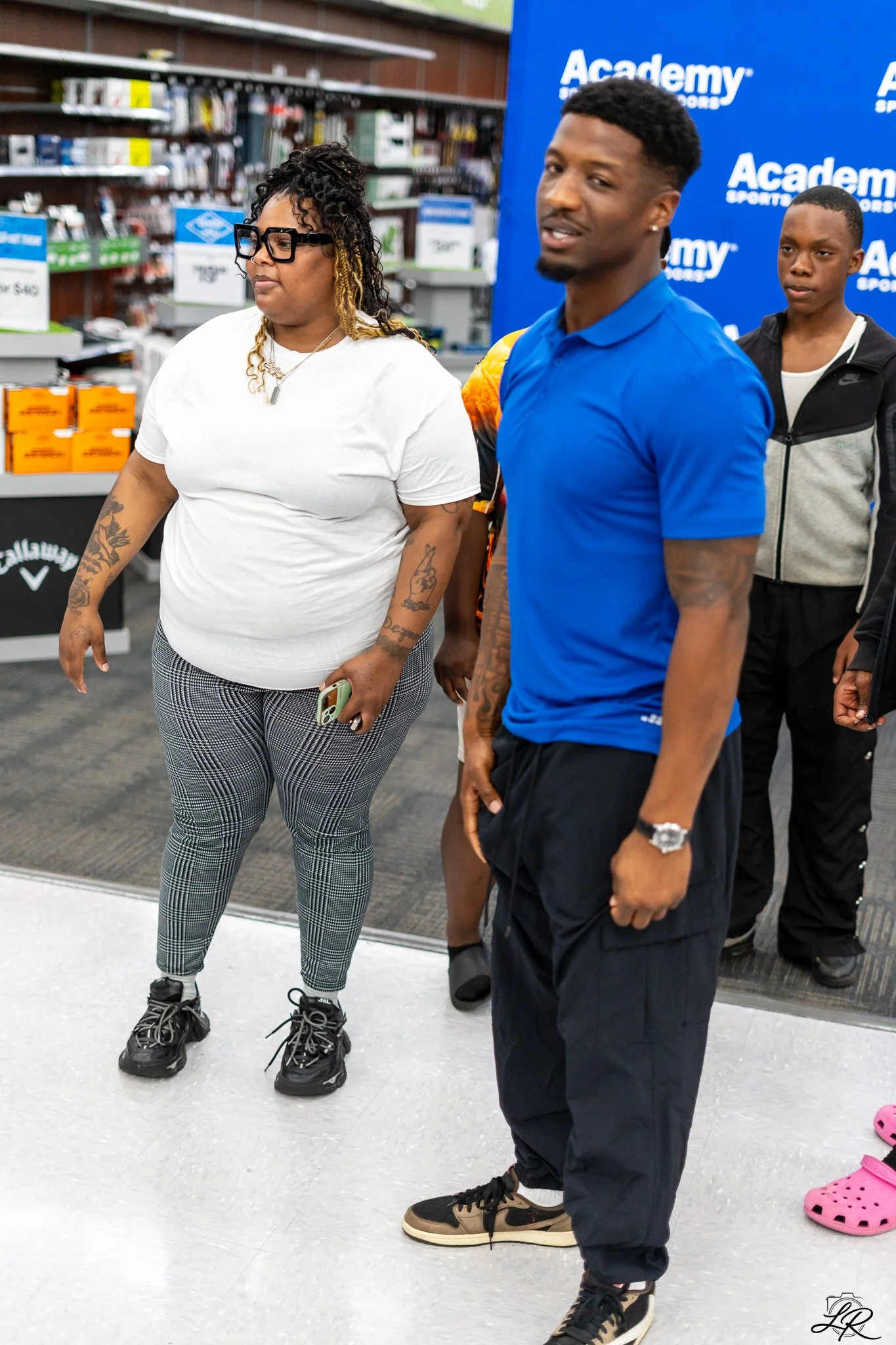 Three people standing inside a store near a blue Academy Sports + Outdoors background. The woman on the left has longer black hair with blonde curls, glasses, tattoos on her arms, and is wearing a white T-shirt and checkered pants. The man in the cen