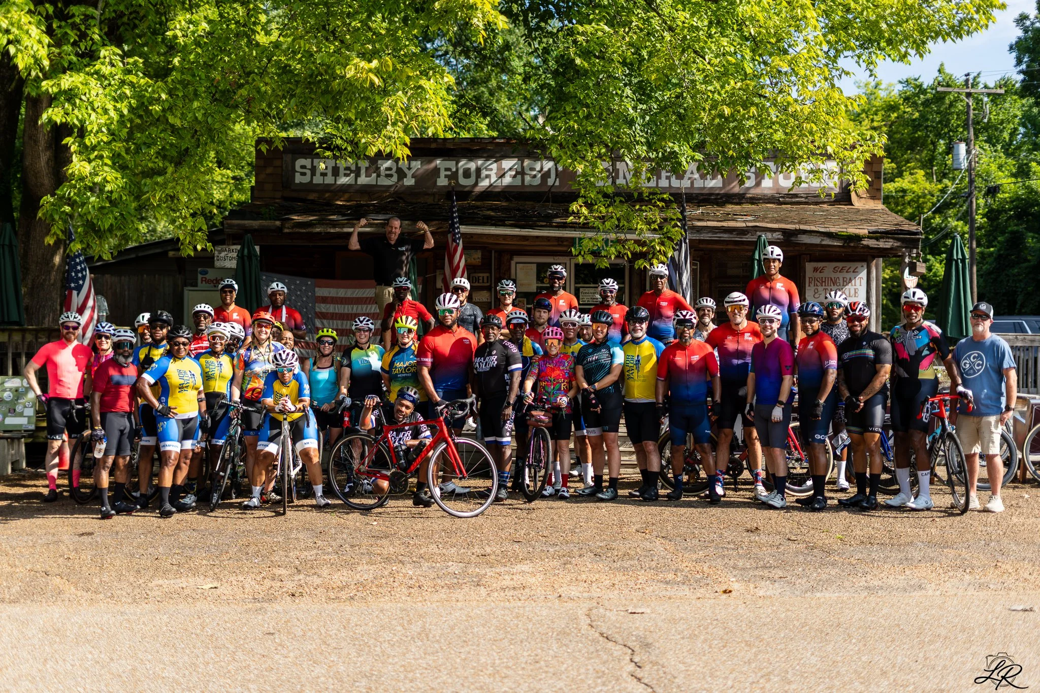 Group of cyclists posing in front of a rustic building with a sign reading 'Shelby Forest, Memphis', surrounded by green trees and American flags.