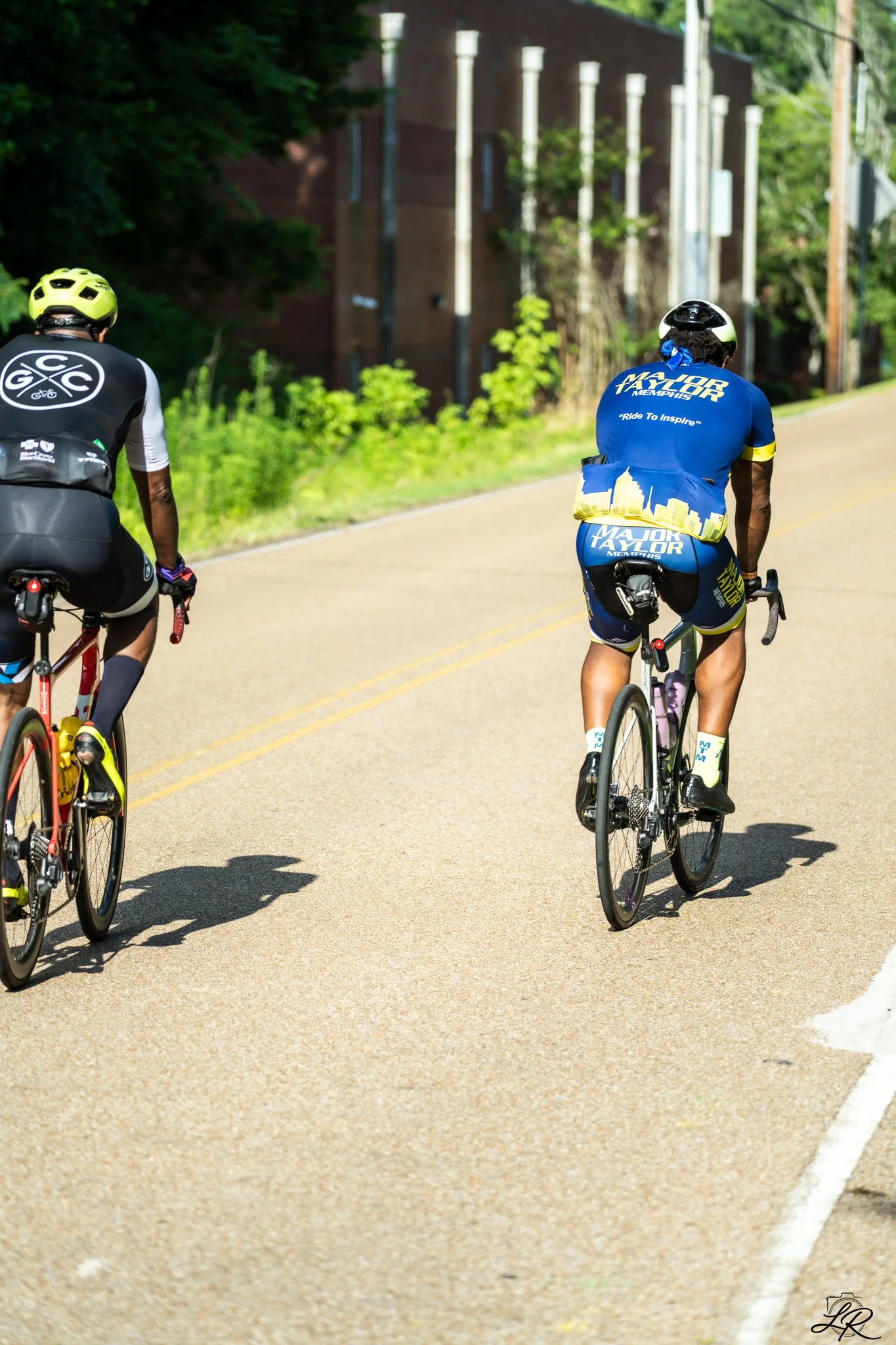 Two cyclists riding on a paved road with trees and a brick building in the background. One cyclist wears a yellow helmet and a black and white jersey; the other wears a white helmet and a blue jersey with yellow accents.
