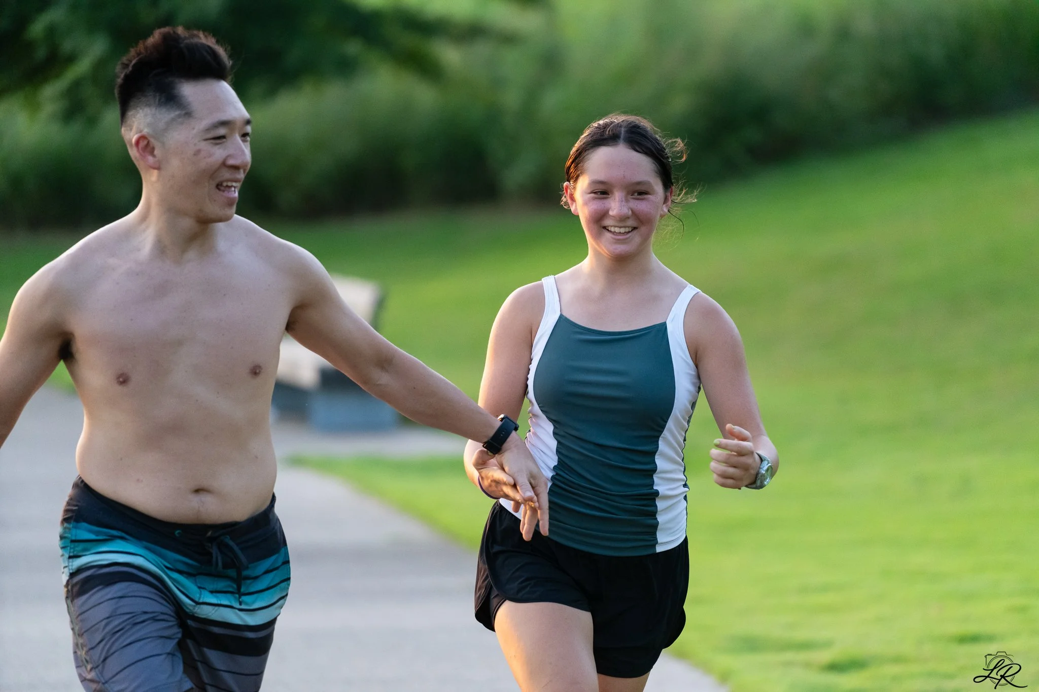 A shirtless man and a woman in a blue athletic tank top and black shorts running outdoors on a grassy trail.