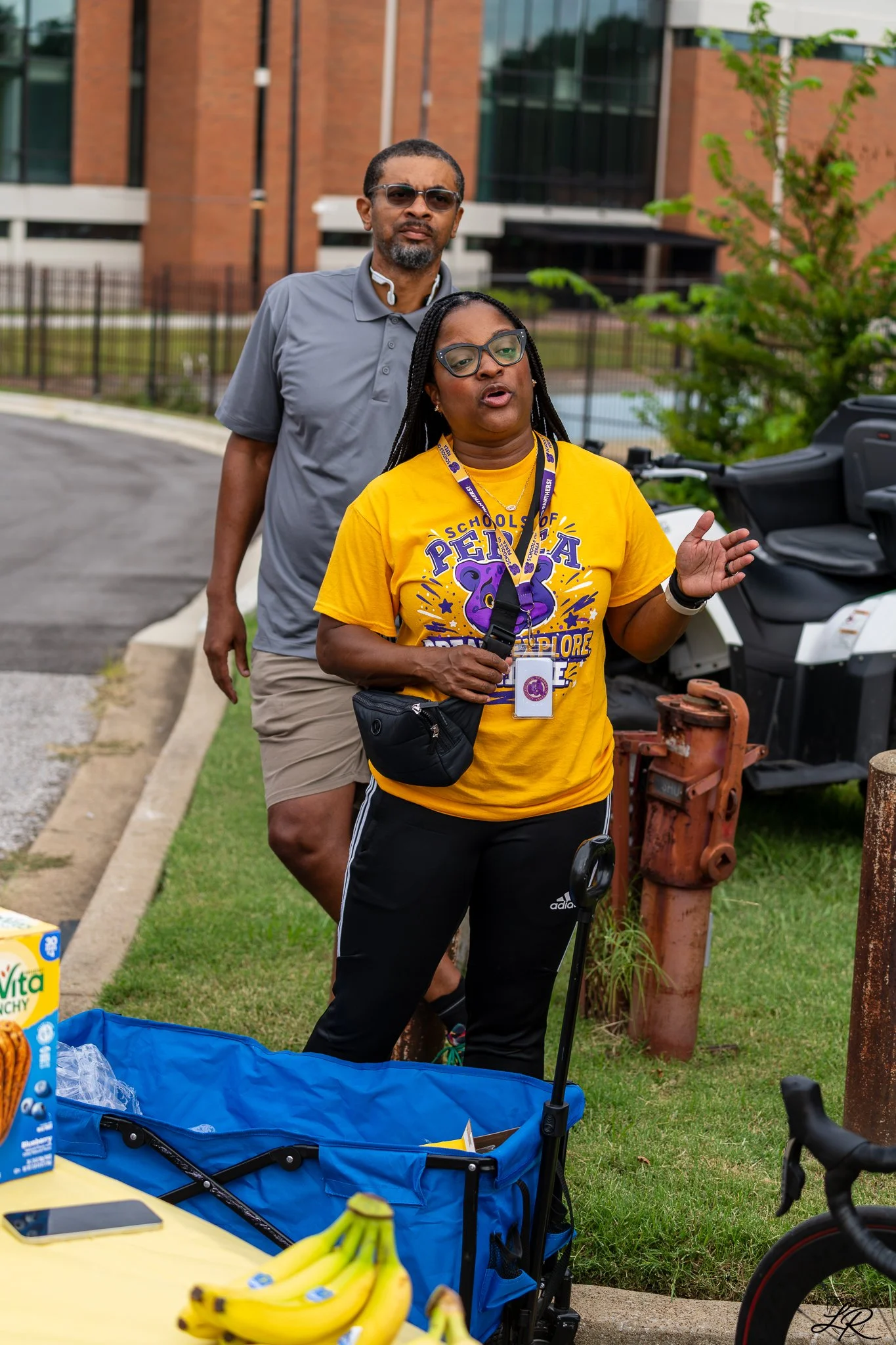 A woman wearing a yellow t-shirt and black Adidas pants, speaking and gesturing outdoors near a fence and trees, with a man standing behind her wearing sunglasses, a gray polo shirt, and beige shorts. There is a blue wagon filled with items, bananas,