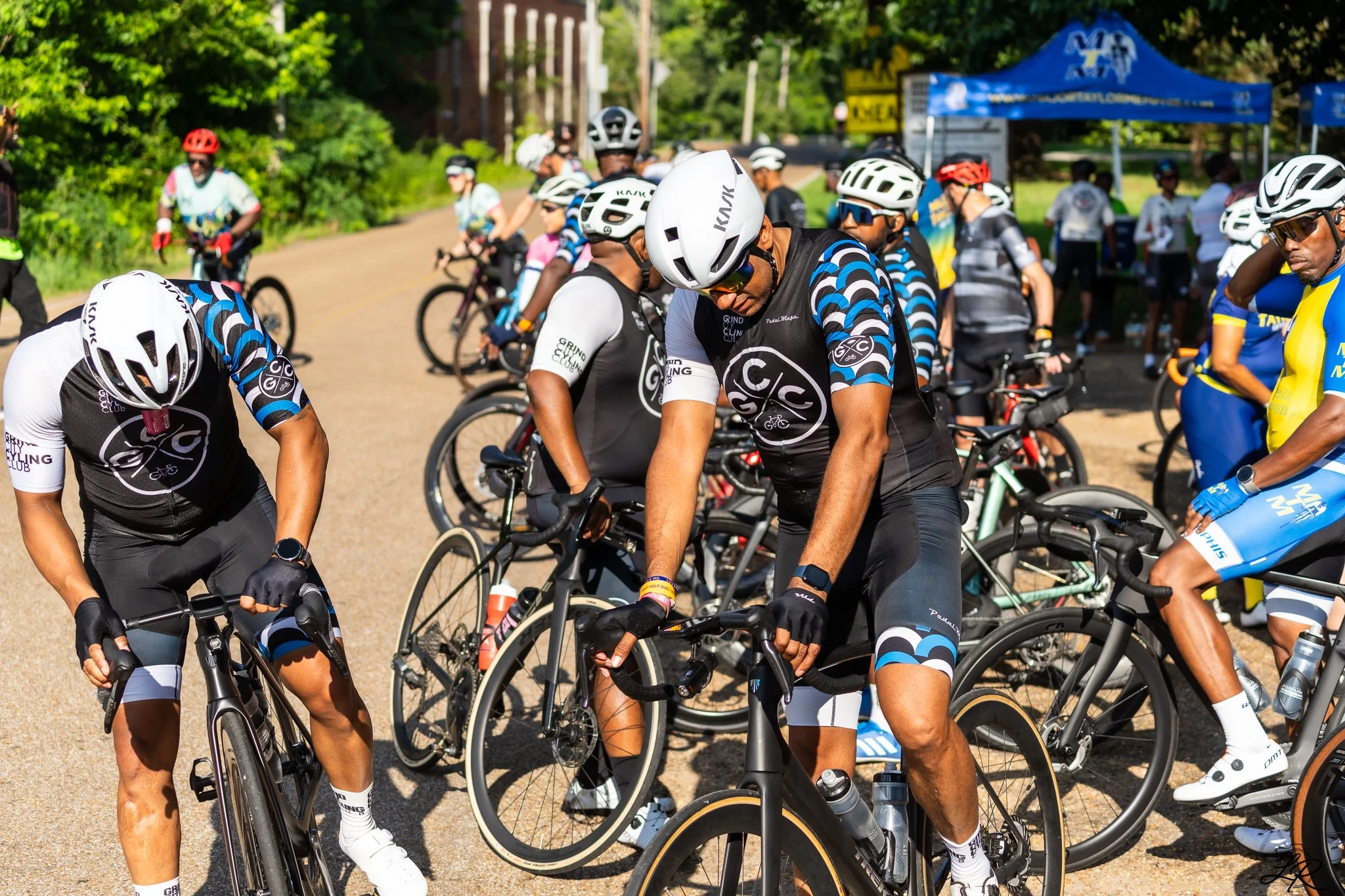 Group of cyclists wearing helmets and cycling gear gathering outdoors, some chatting, with a blue tent in the background and lush green trees.