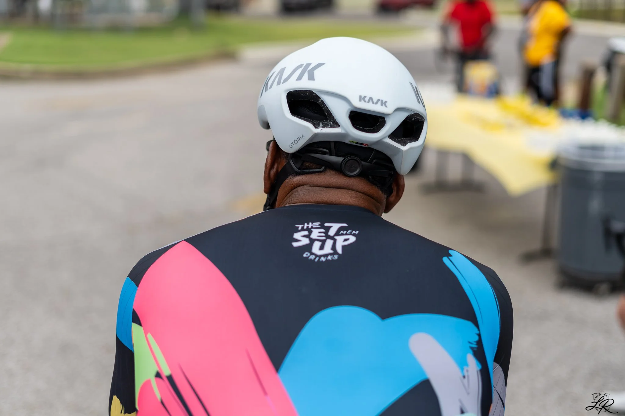 Back of a cyclist wearing a colorful jersey and a white helmet with black vents, with blurred people and tables in the background.