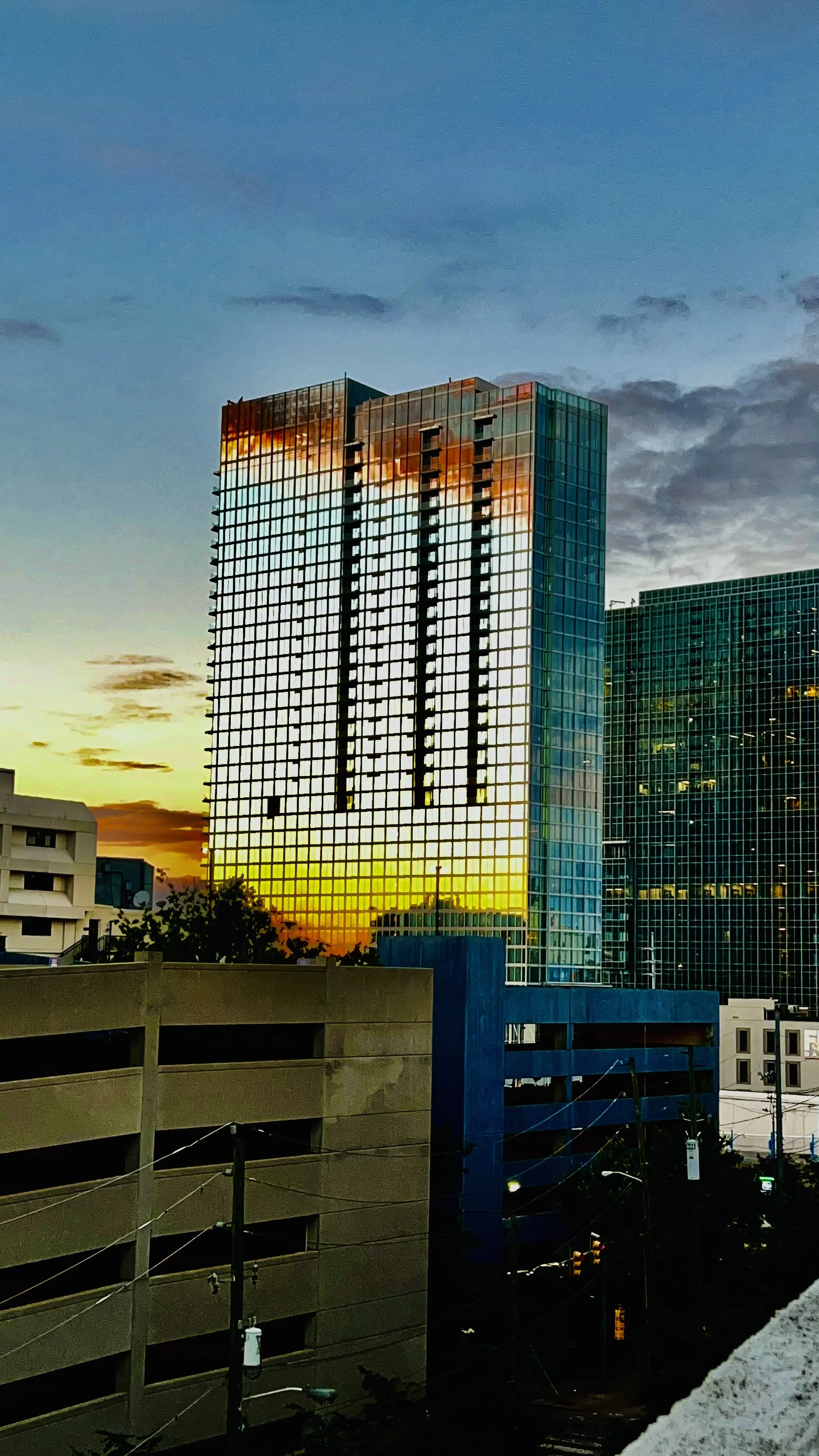 Tall glass building reflecting the sunset sky with clouds during dusk, neighboring buildings and power lines in the foreground.