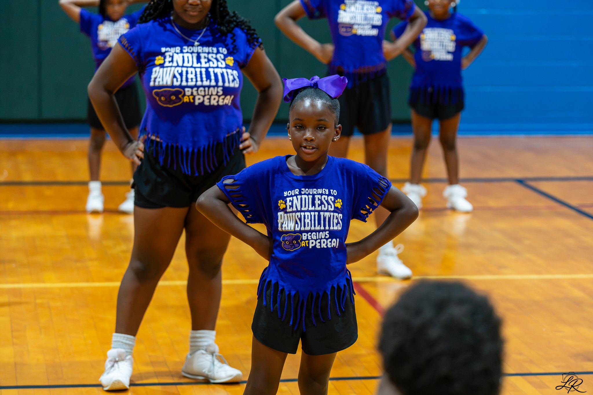 Young girl with a purple bow in her hair standing proud with hands on her hips in a gymnasium, wearing a blue t-shirt with the phrase "Your journey to endless paw-sibilities begins at P.E.R.E.A!" and black shorts, surrounded by her cheerleading teamm