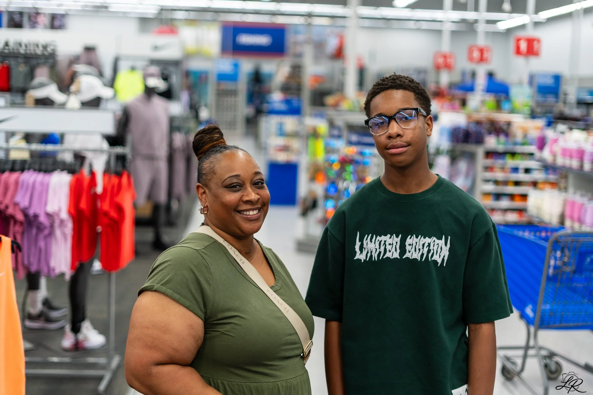 A woman and a young man standing inside a retail store, smiling at the camera, with clothing and other merchandise visible in the background.