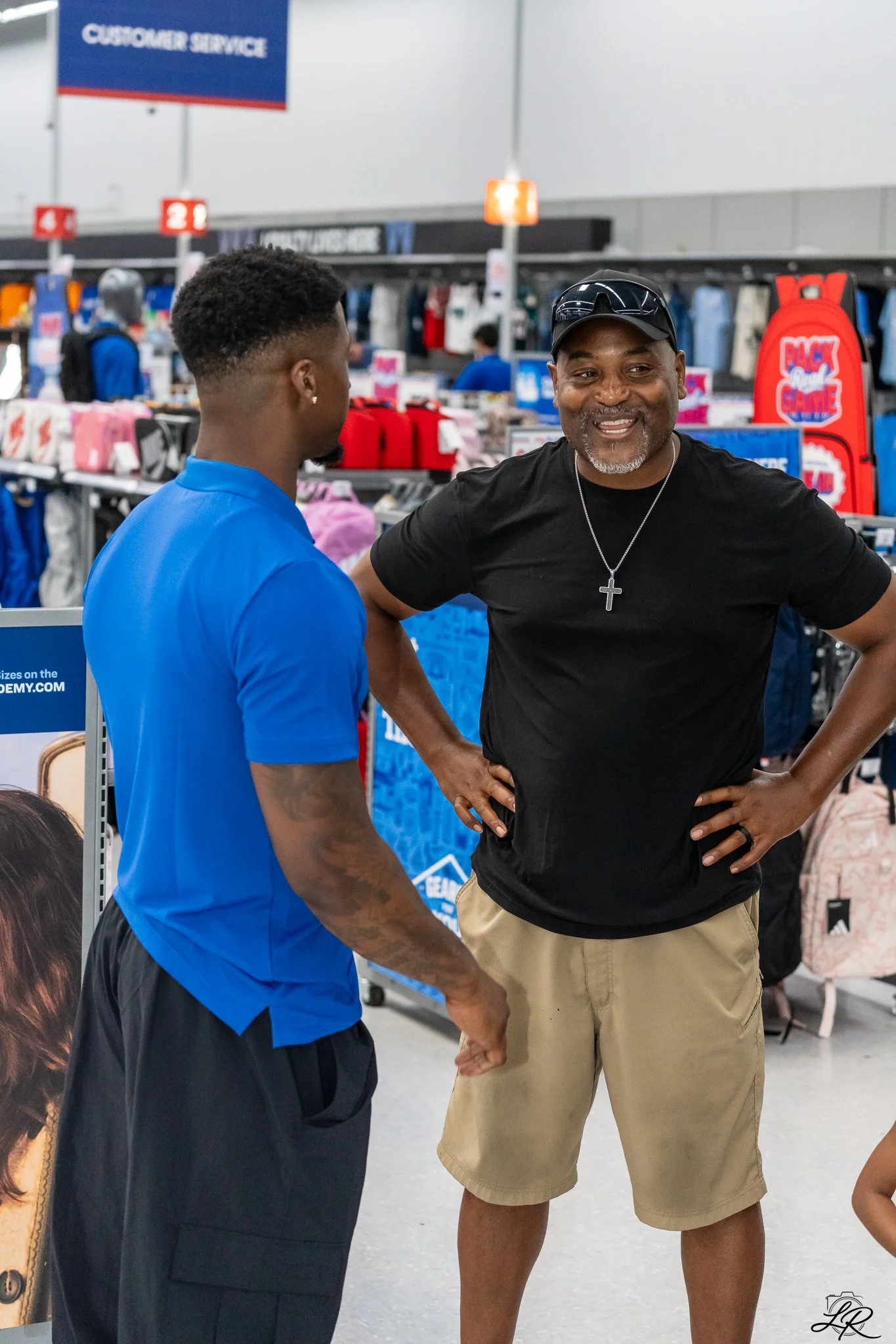 Two men are talking at a store checkout counter, one wearing a blue shirt and the other in a black shirt and cap, with a customer service sign visible in the background.